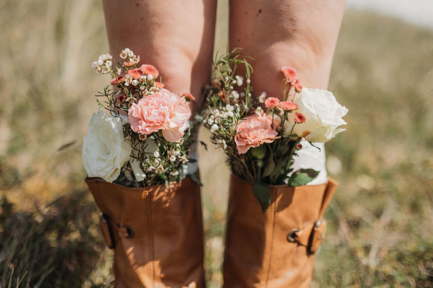 Brown leather boots filled with pink and white carnations and wildflowers on a grassy field.