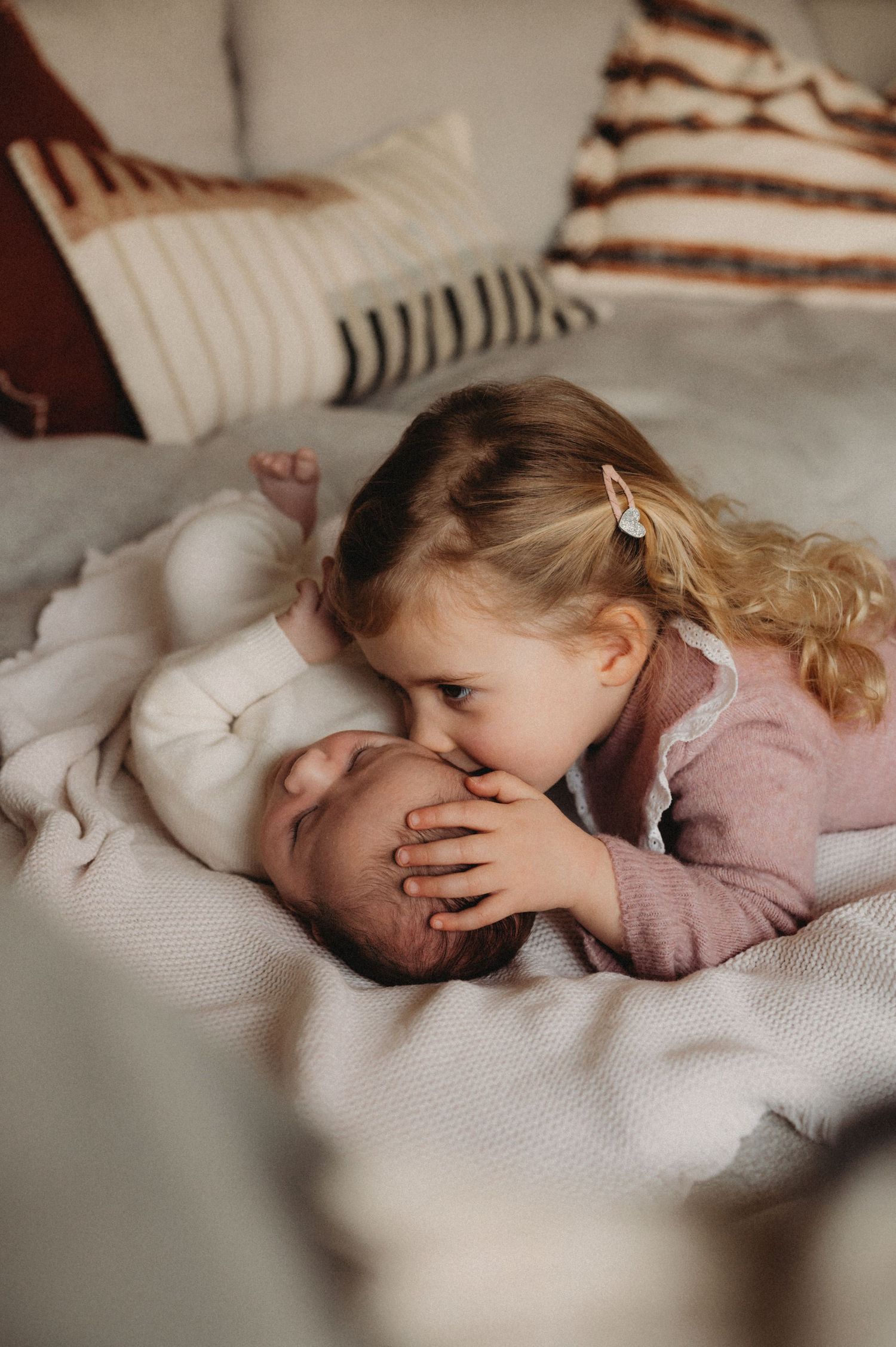 A young blonde girl lovingly kisses her newborn sibling on a cozy bed with pillows.