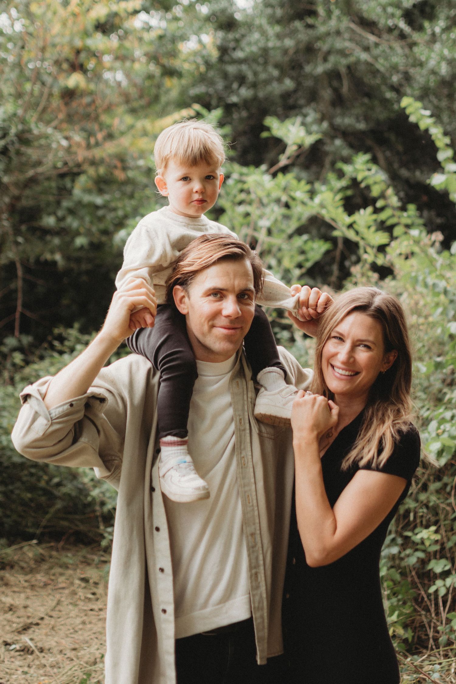 Smiling family with toddler sitting on fathers shoulders outdoors in a lush green nature setting.