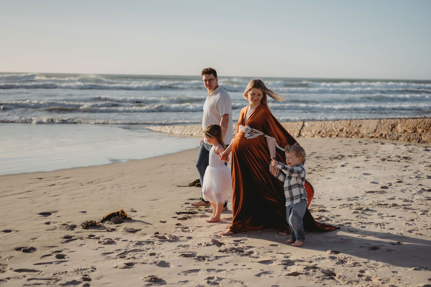 A young family with two toddlers poses together on a sandy beach at golden hour.