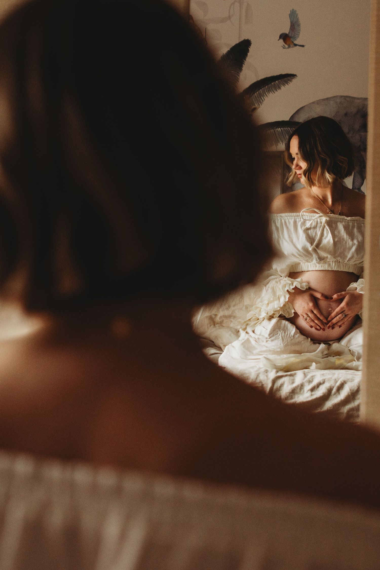 Pregnant woman in white crop top making heart shape with hands on baby bump, reflected in mirror.