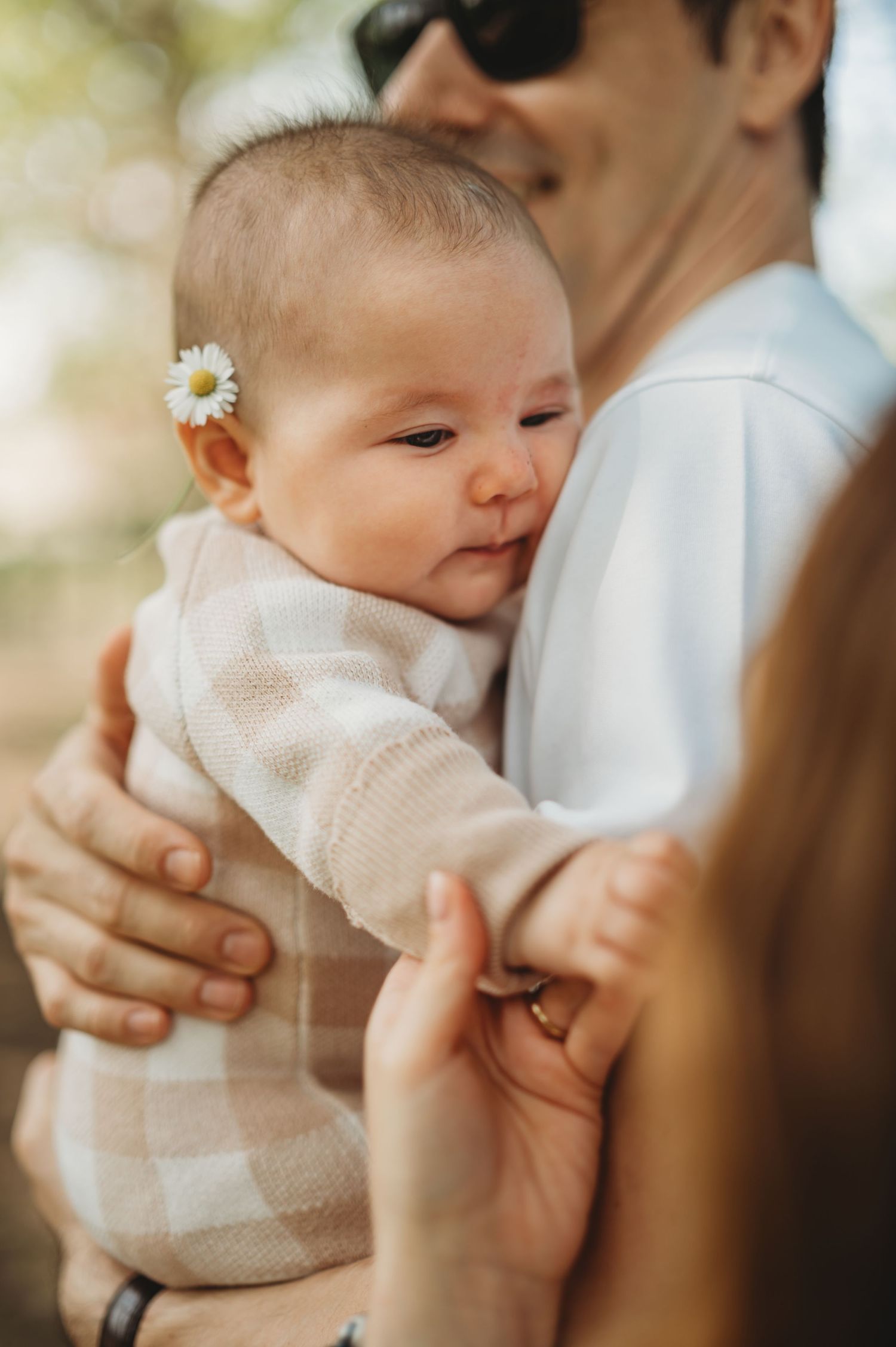 A father holds his newborn baby girl with a daisy flower in her hair during an outdoor family photo session.