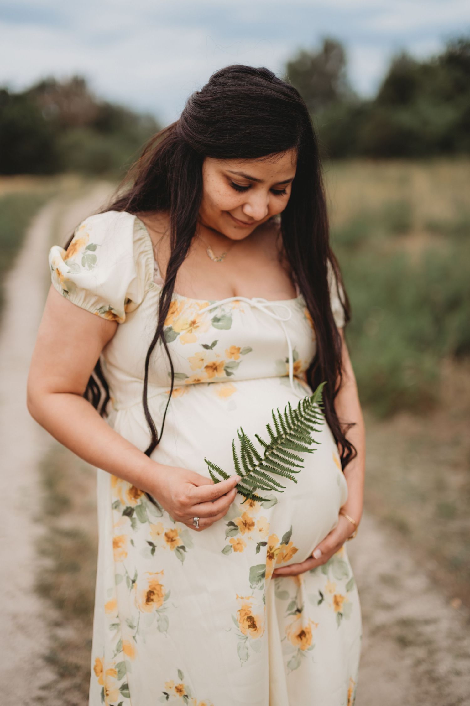 Smiling pregnant woman in floral dress holding fern leaf against baby bump.