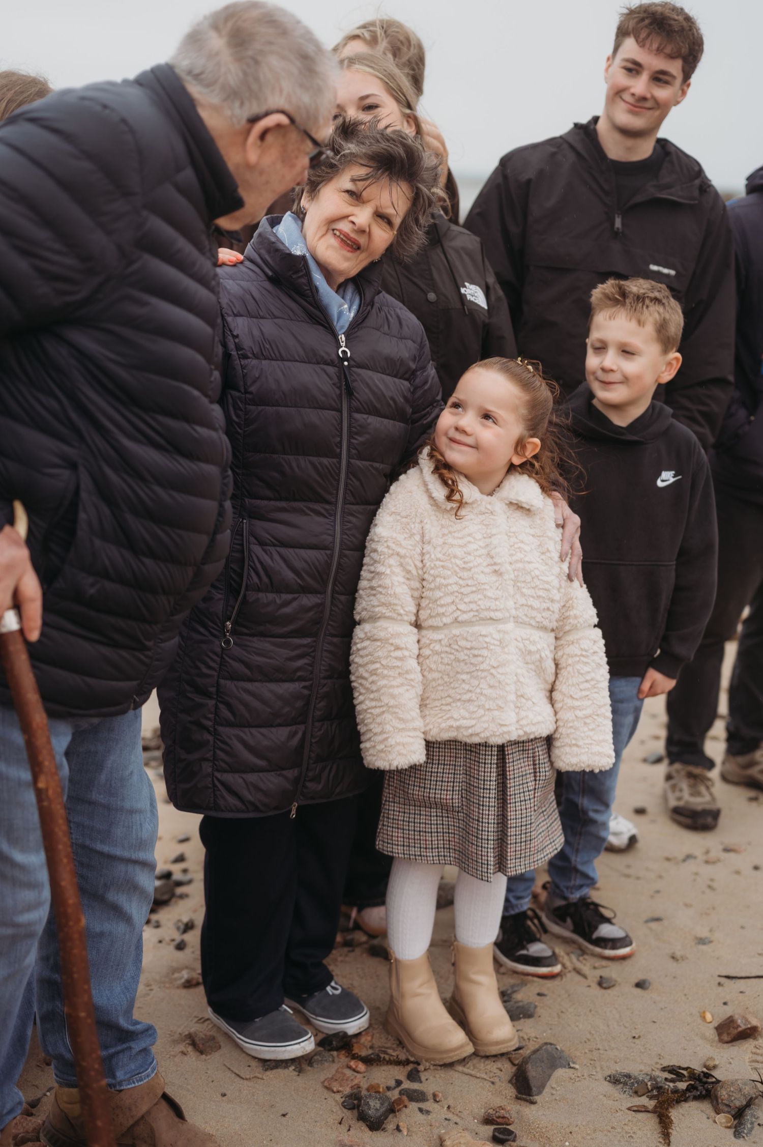 Young girl in cream coat smiling with family group on a sandy beach on a cloudy day.