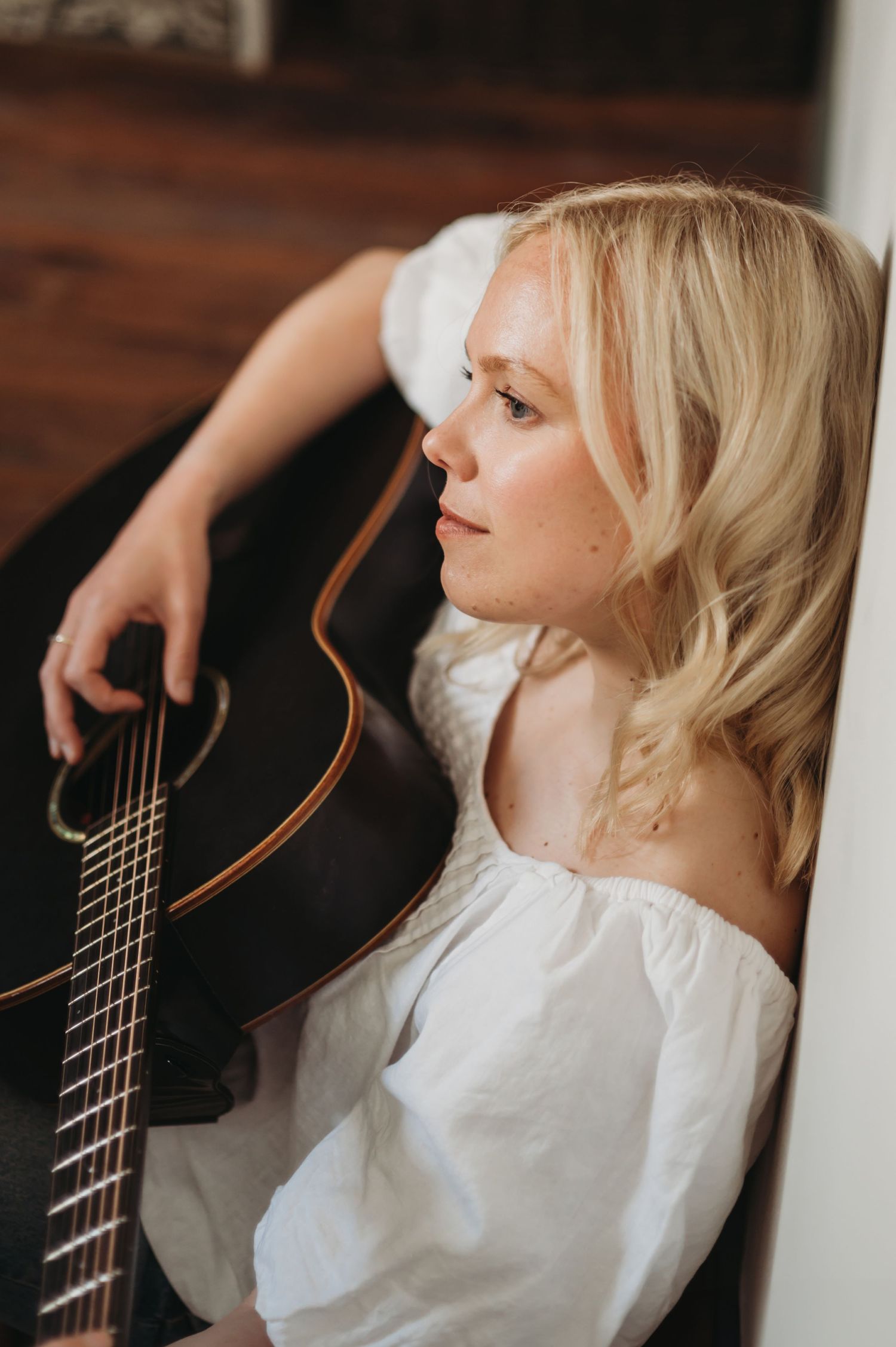 Blonde woman in white off-shoulder top holding acoustic guitar while sitting against a wall.