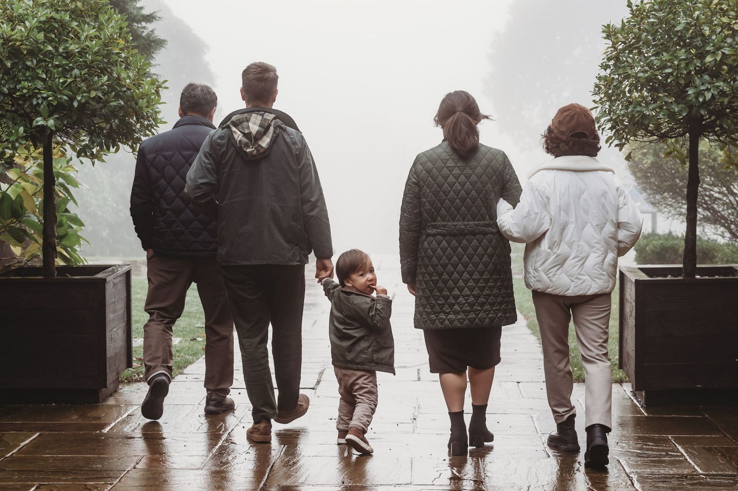 Family of five walking away on a rainy foggy day, parents and grandparents holding hands with a toddler.
