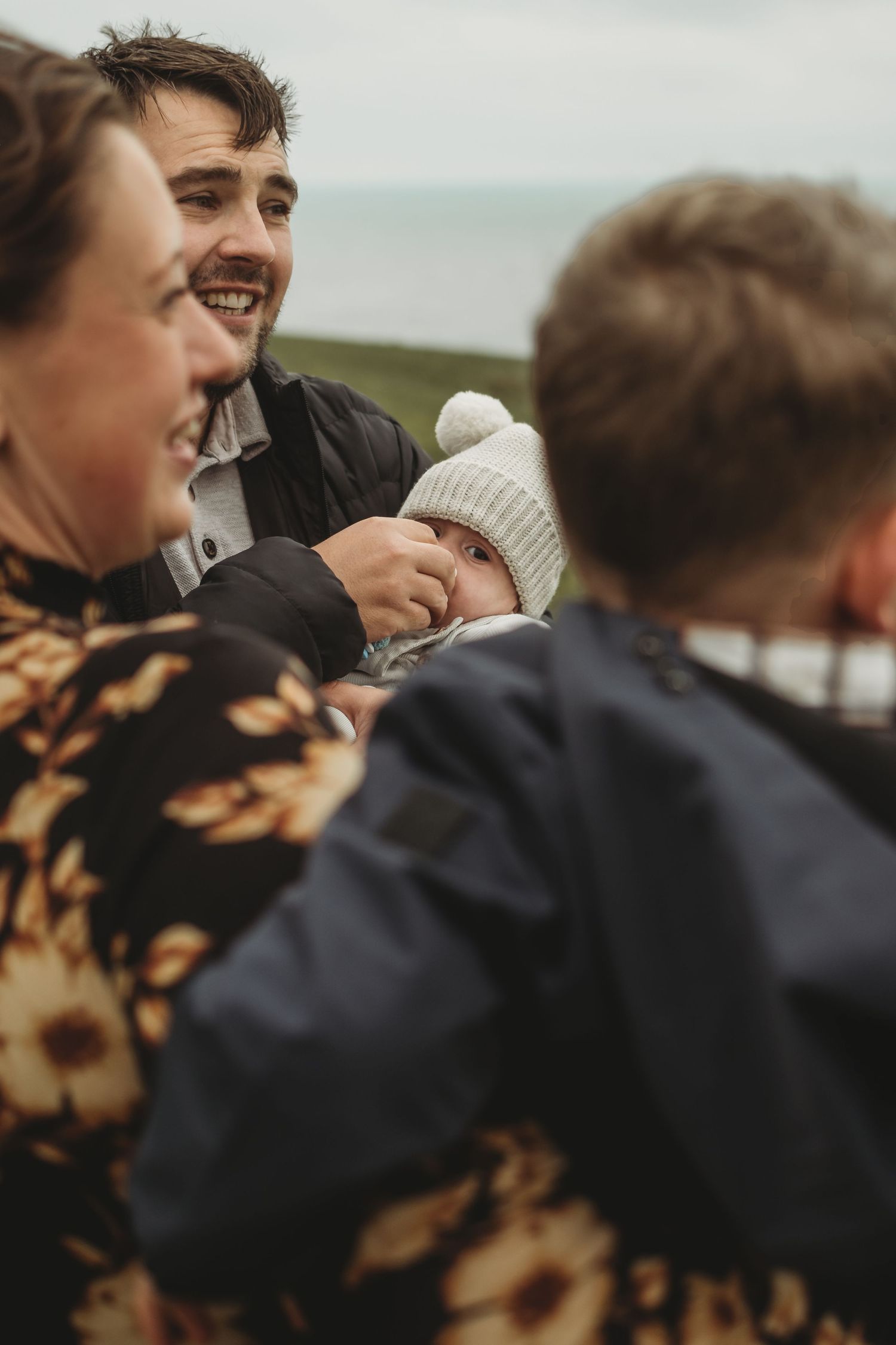 Smiling family with a baby in a knit hat gathered together outdoors near the coast.