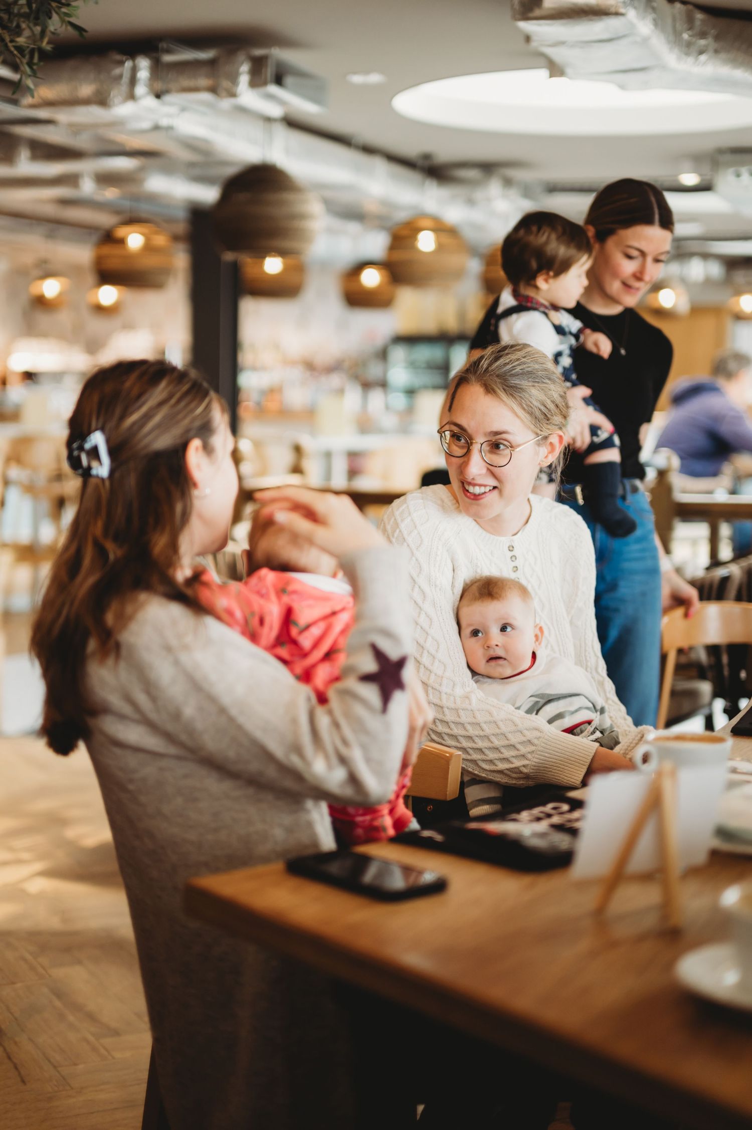 Mothers with babies chatting and smiling at a warm indoor parenting group meetup event.