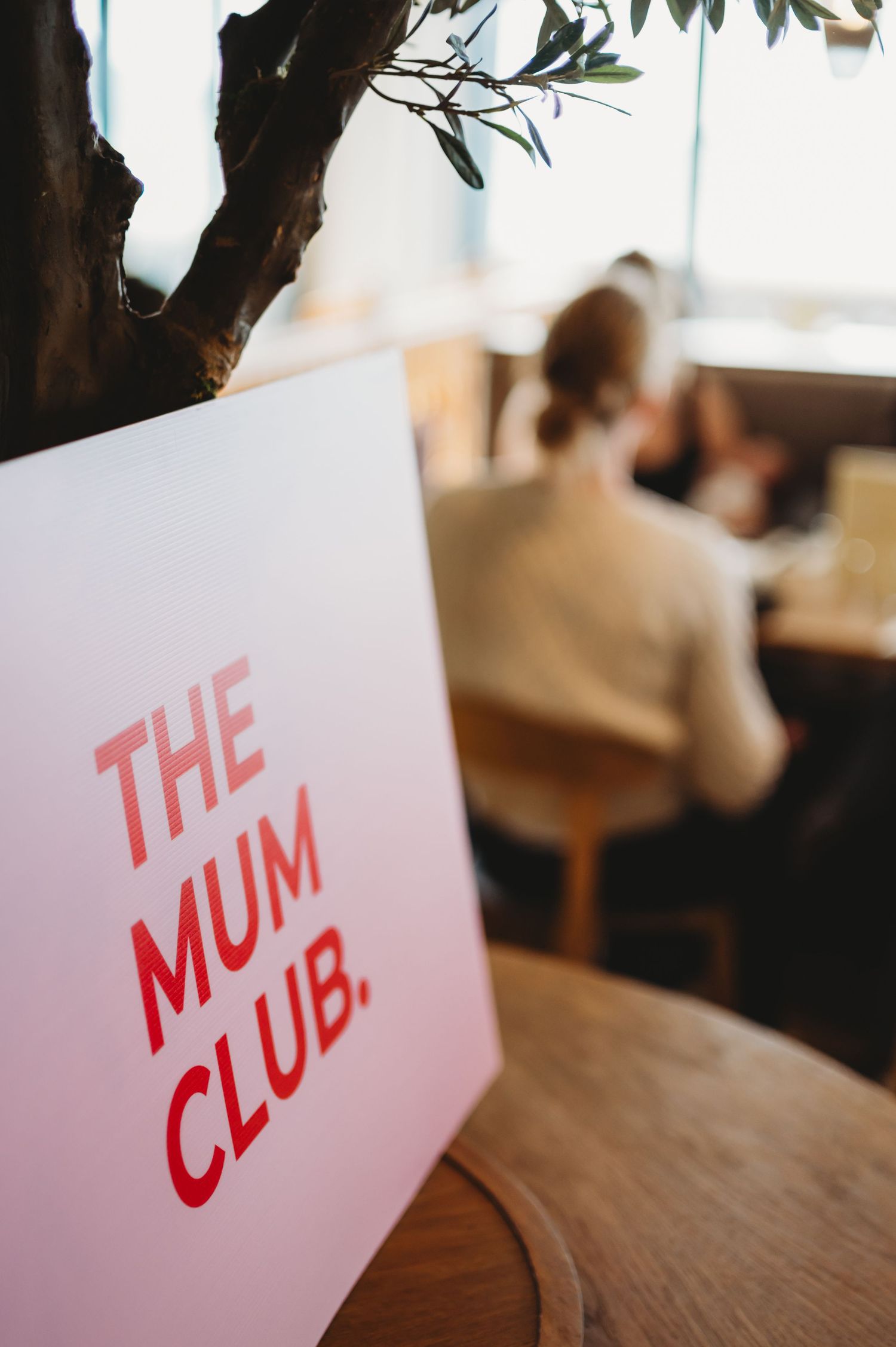 The Mum Club pink sign displayed on a wooden table in a cozy cafe setting.