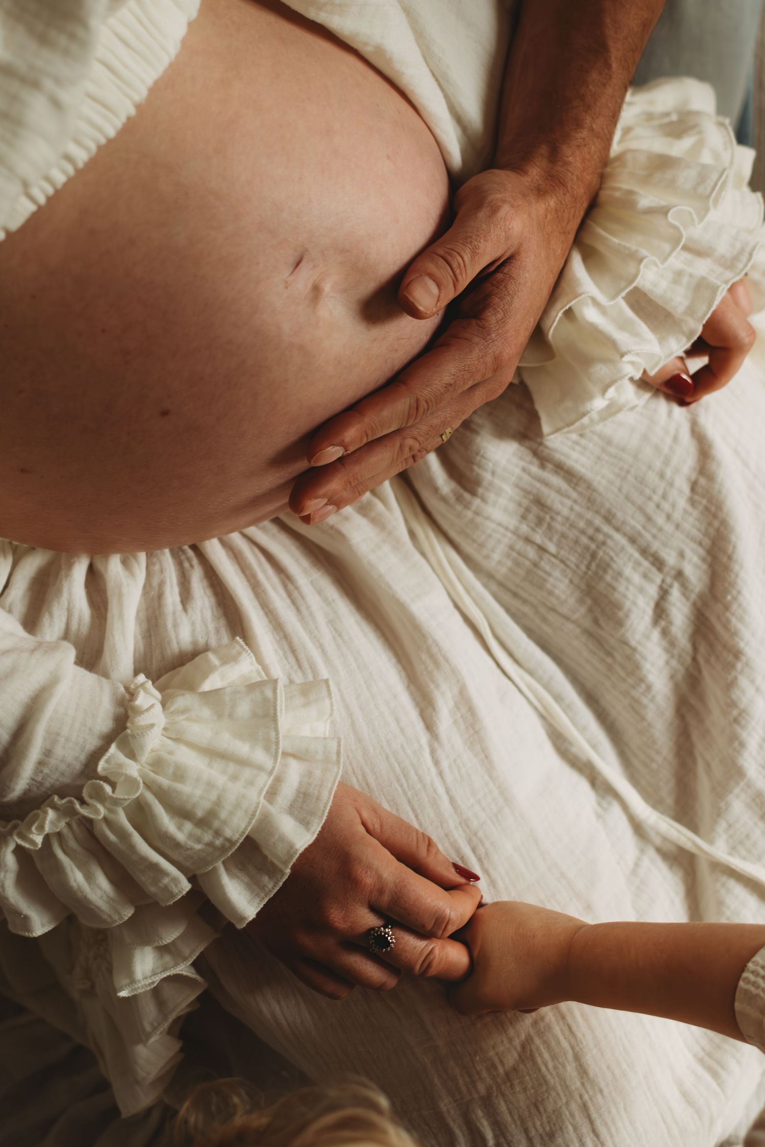Close-up of pregnant belly in white ruffled dress with partners hand and childs hand touching it.