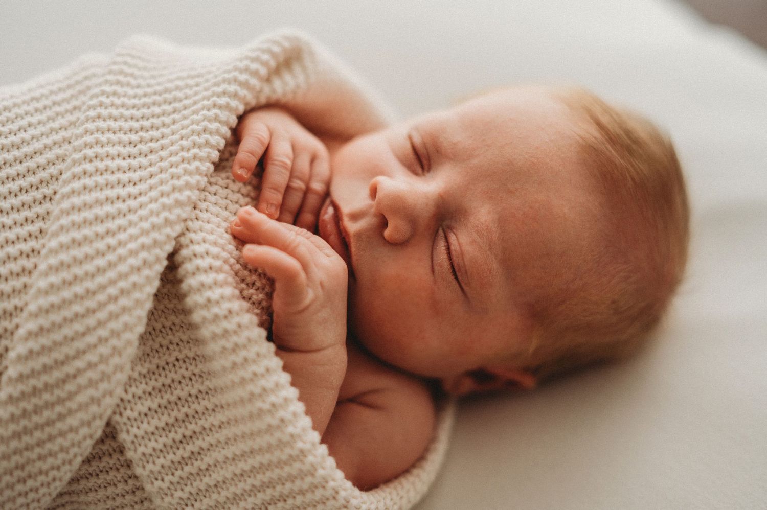 Peaceful sleeping newborn baby wrapped in a soft beige knit blanket on a white surface.