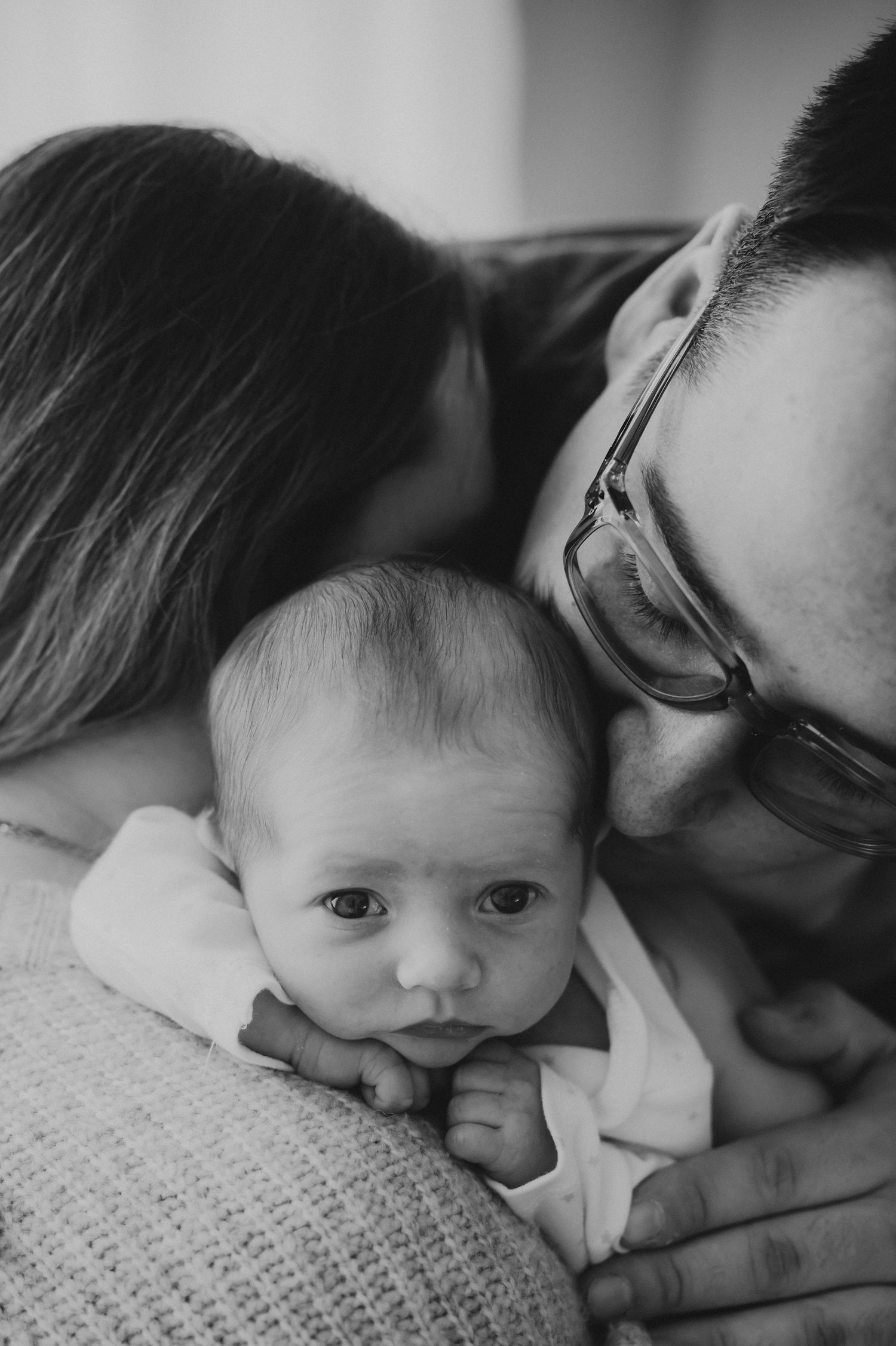 Black and white photo of parents lovingly embracing their alert newborn baby.