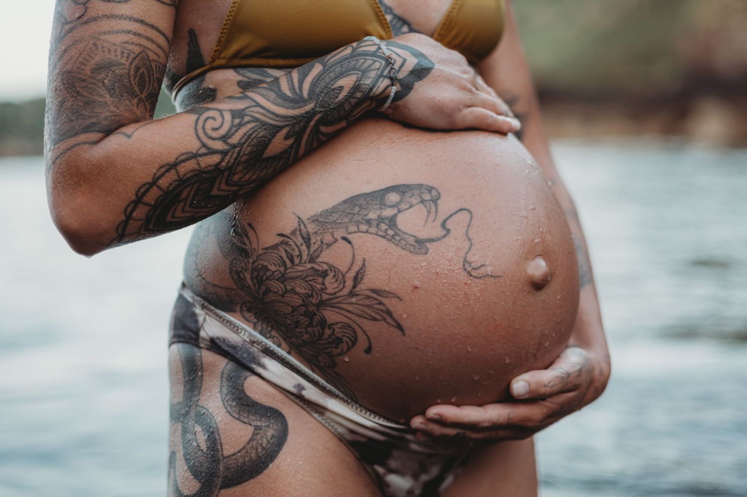 Pregnant woman with intricate snake and floral tattoos cradling her baby bump near water.