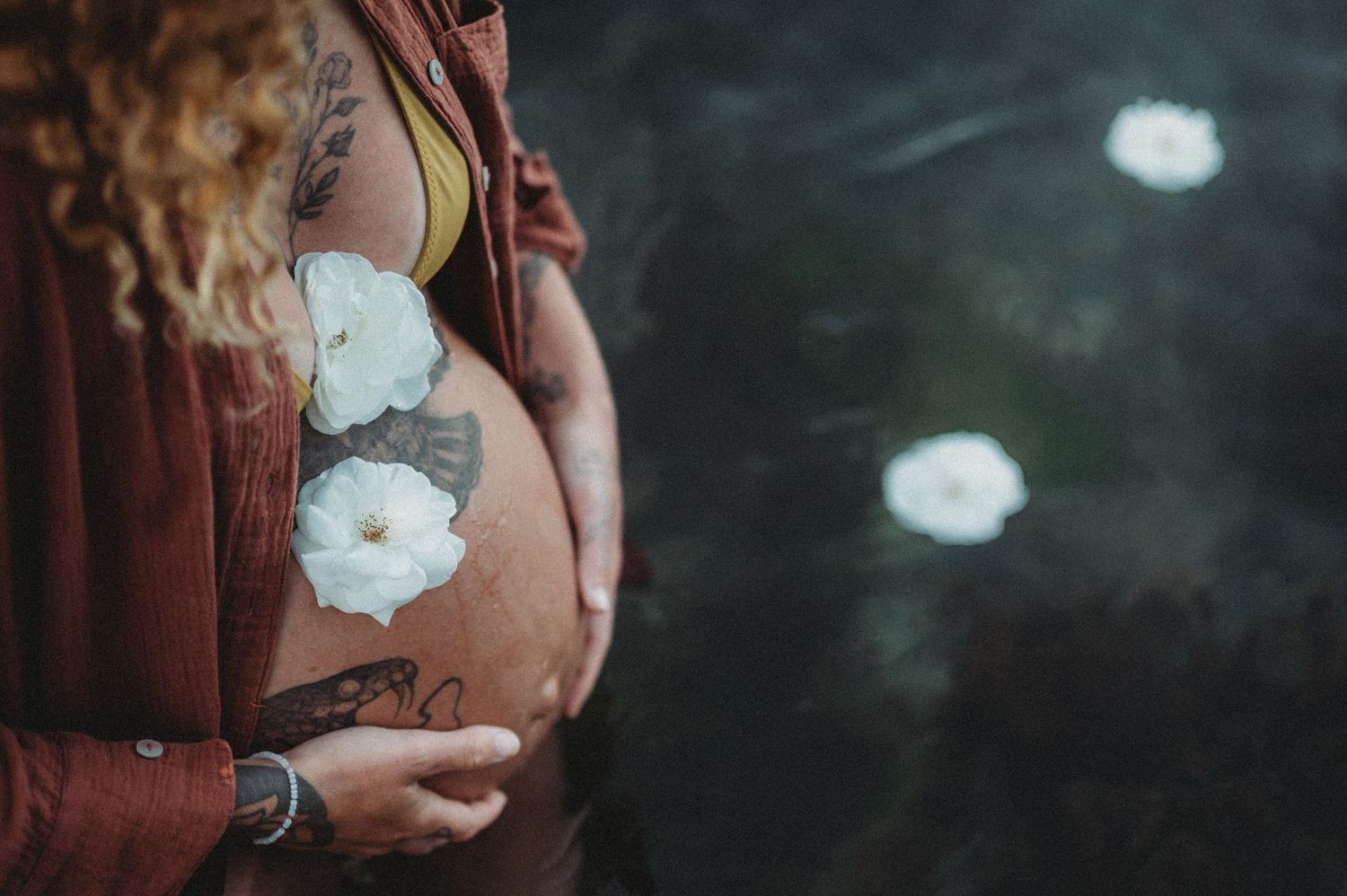 Pregnant woman with tattoos and white flowers on her bare baby bump, wearing a rust-colored jacket outdoors.
