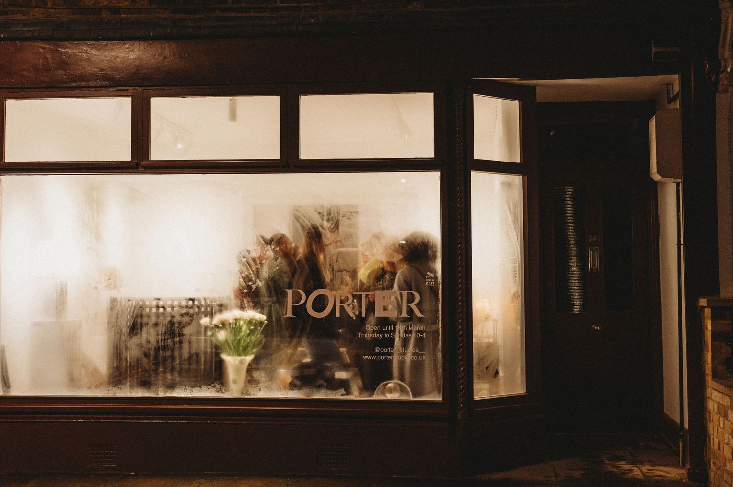 Warmly lit PORTER studio storefront window with dried flowers at night.
