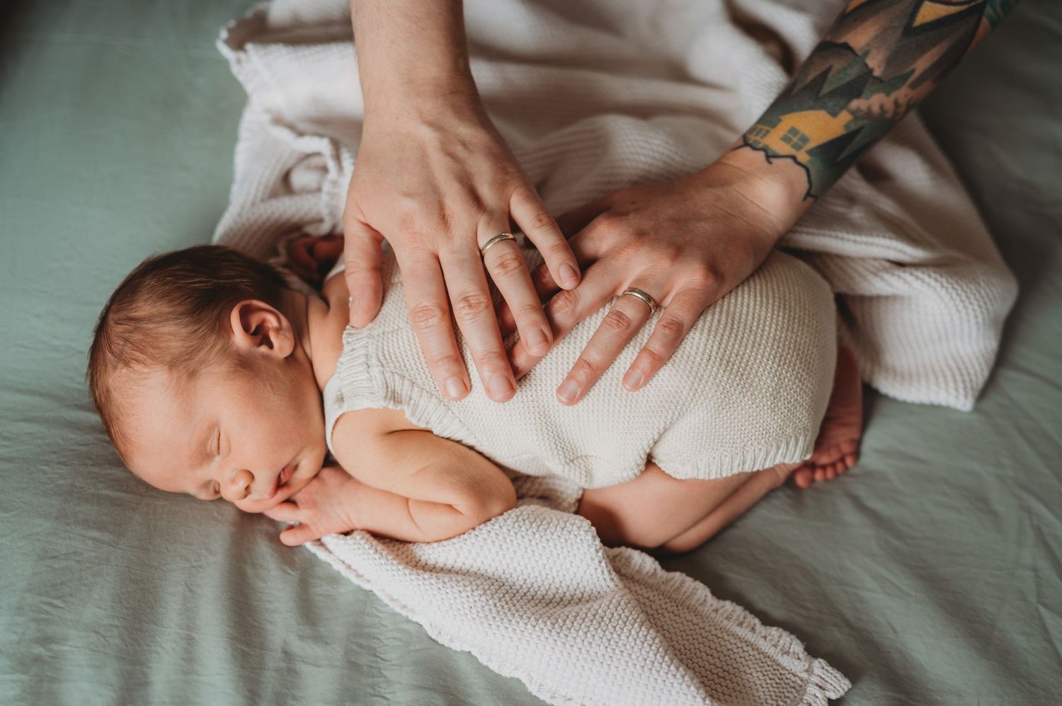 Parent with tattoos gently holding a sleeping newborn baby on a gray blanket.