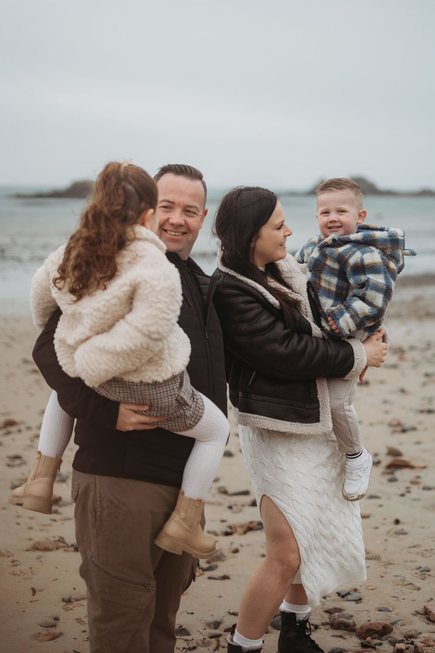 Happy family of four on a beach, parents each holding a young child while smiling at each other.
