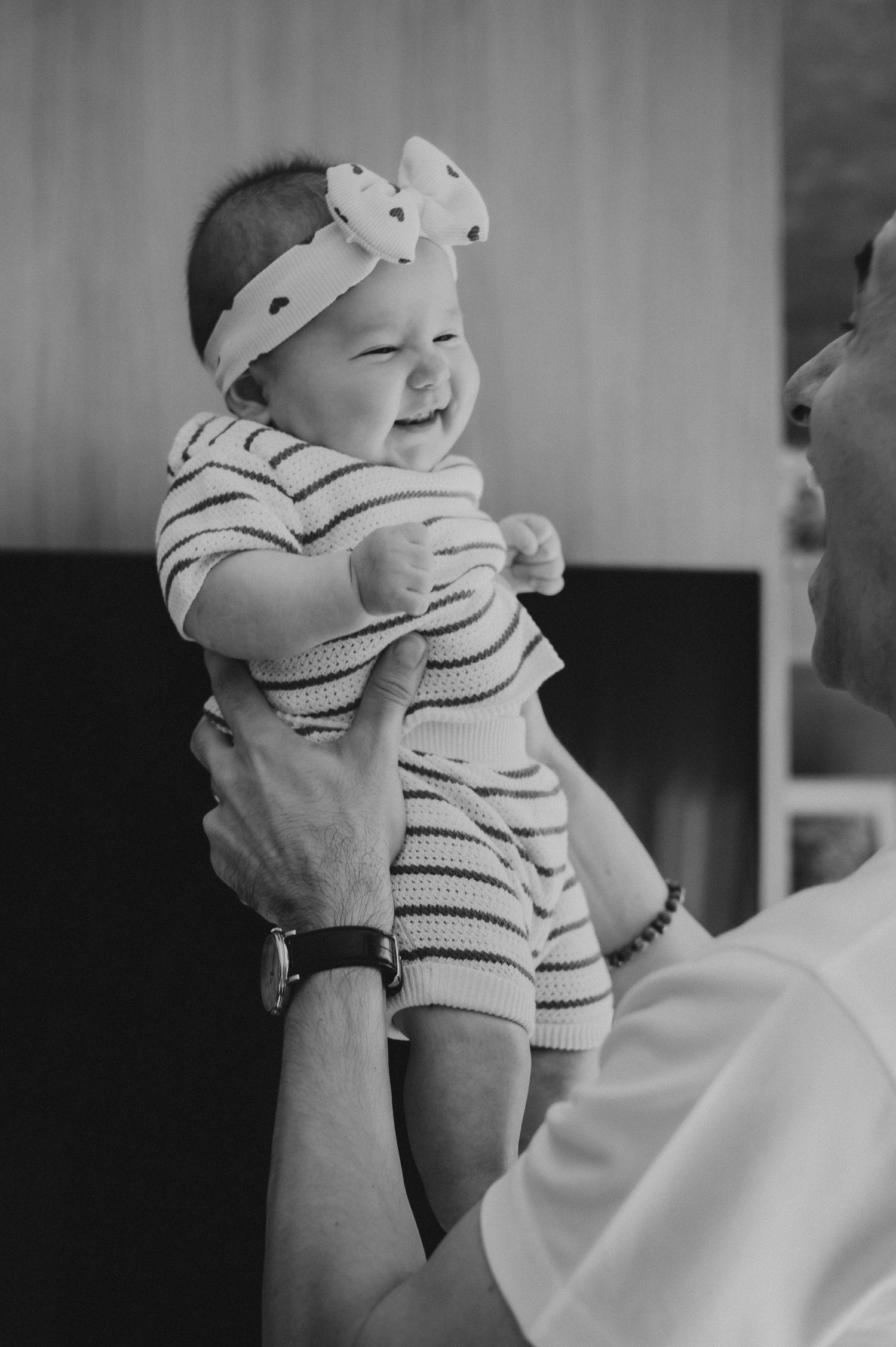 Black and white photo of a laughing baby girl in bow headband being lifted by father.