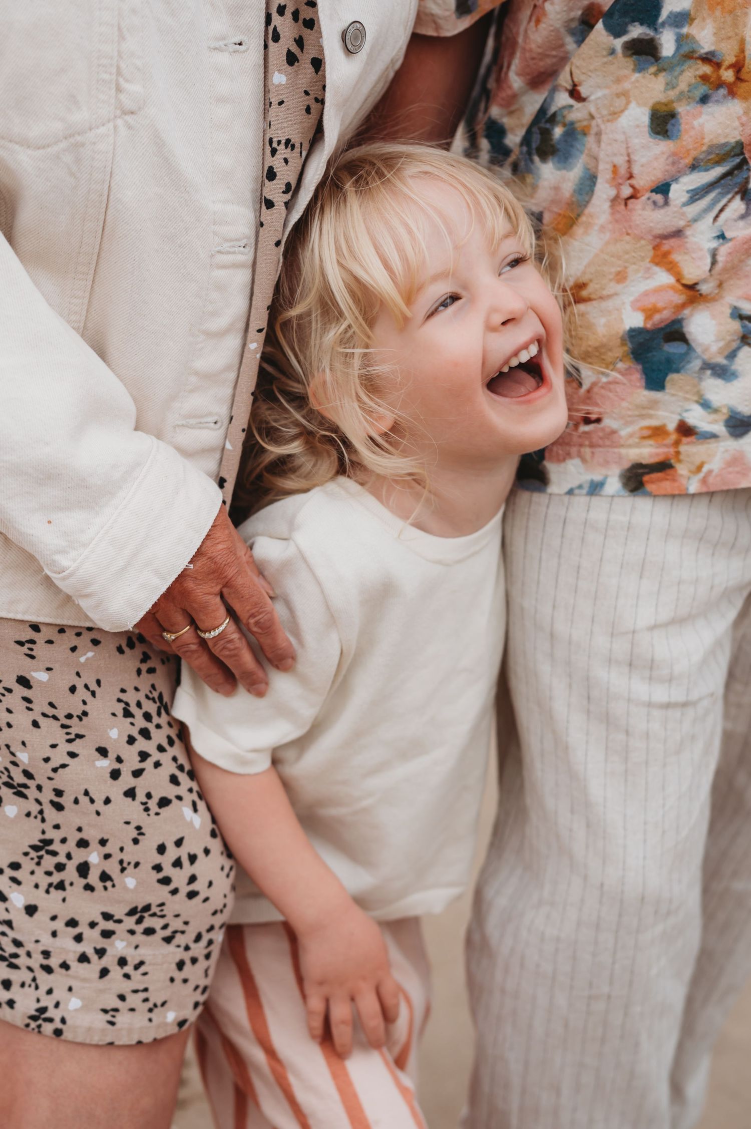 Joyful blonde toddler laughing while standing between two adults in casual neutral clothing.
