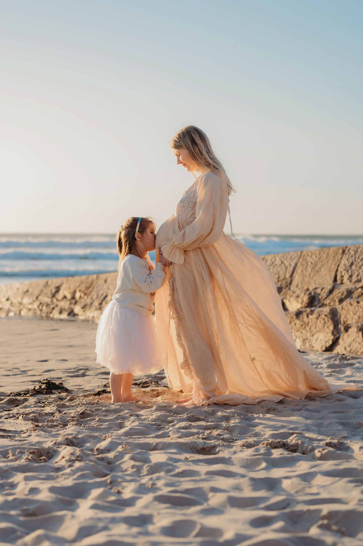 Pregnant woman in peach gown sharing tender moment with young daughter on sandy beach at sunset.