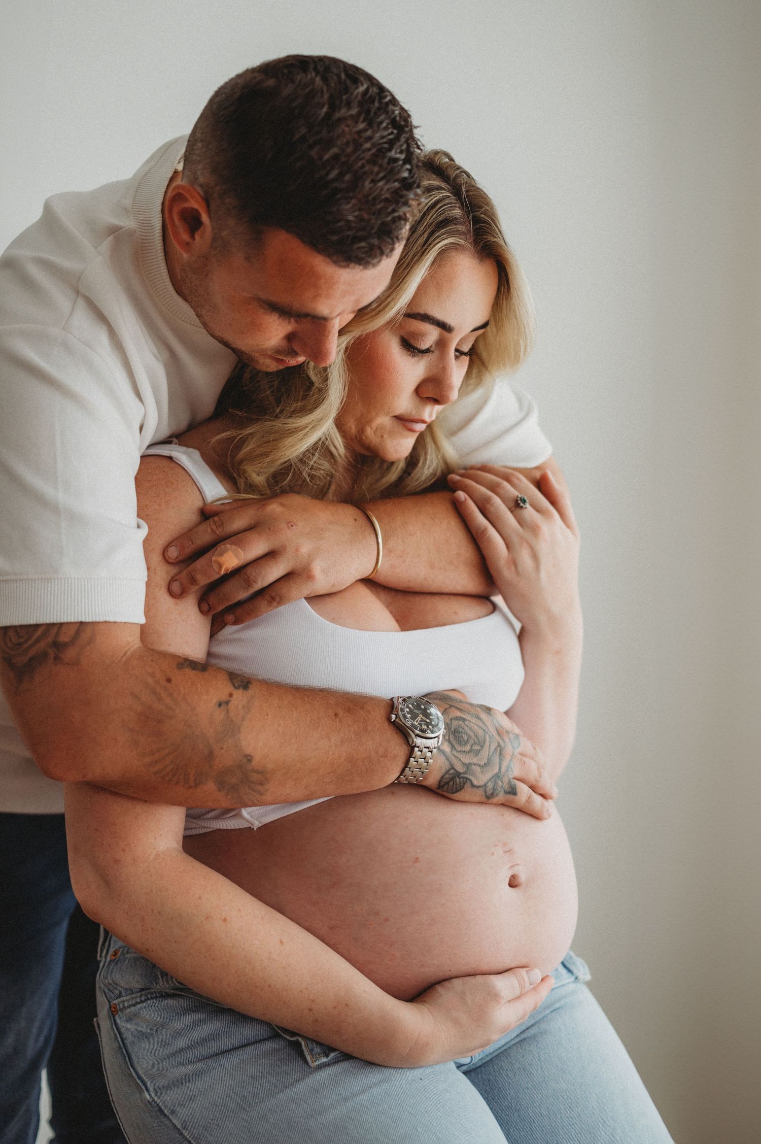 A tattooed man embraces his pregnant partner from behind, both cradling her bare baby bump.