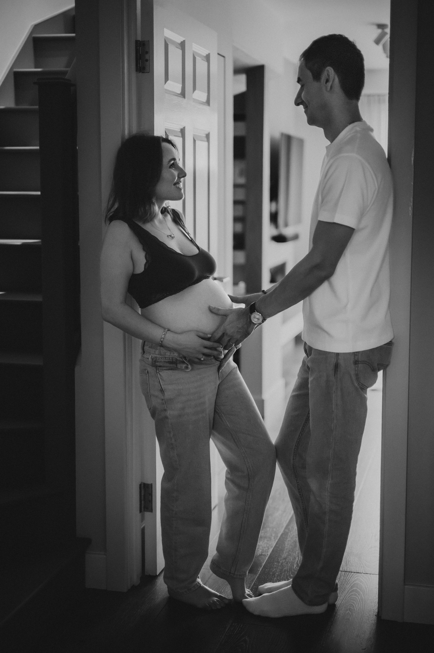 Black and white photo of a couple sharing a tender moment, hands on pregnant belly, standing in a doorway.