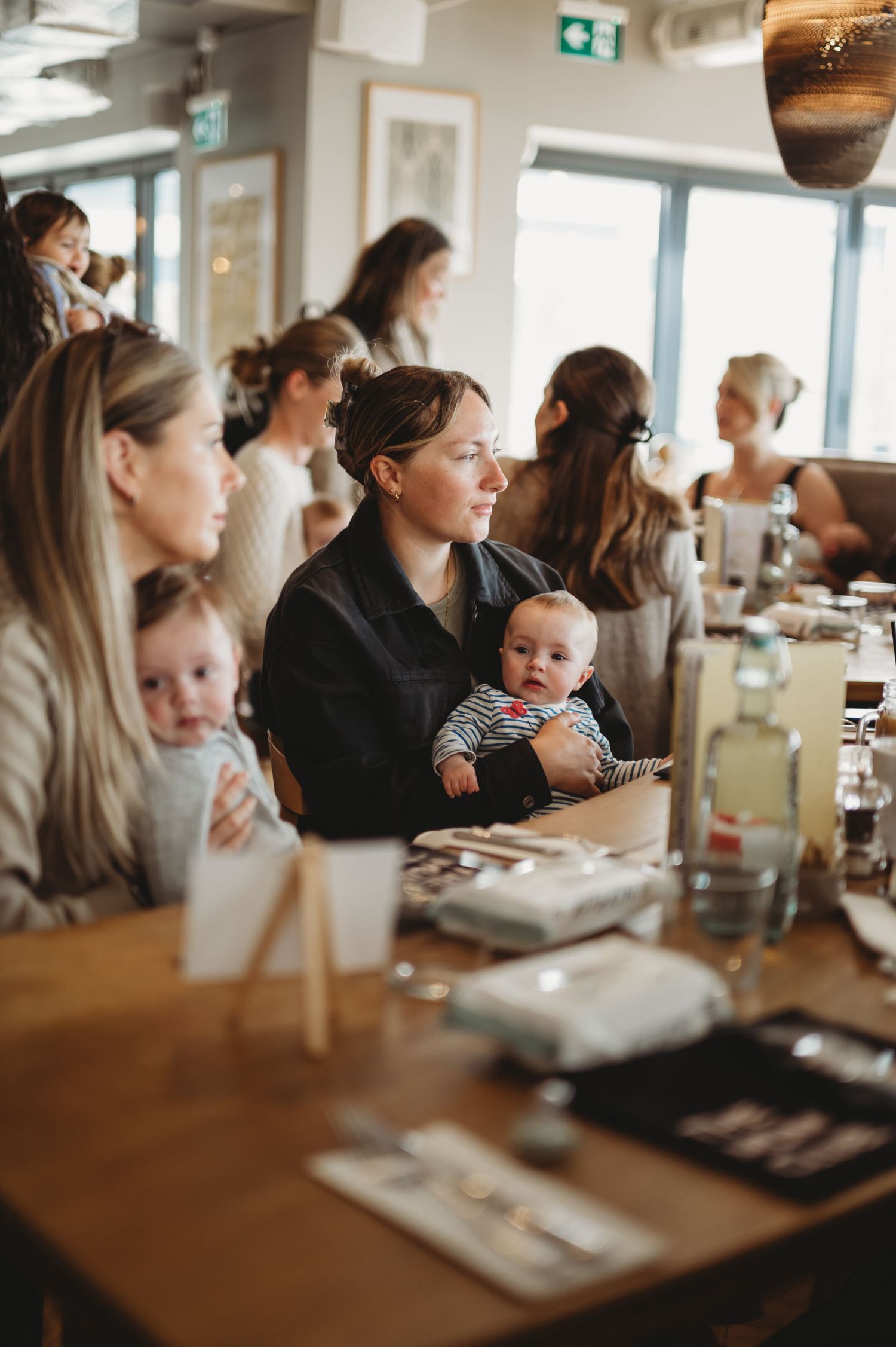 Mothers with babies seated at a long wooden table during an indoor parenting meetup event.