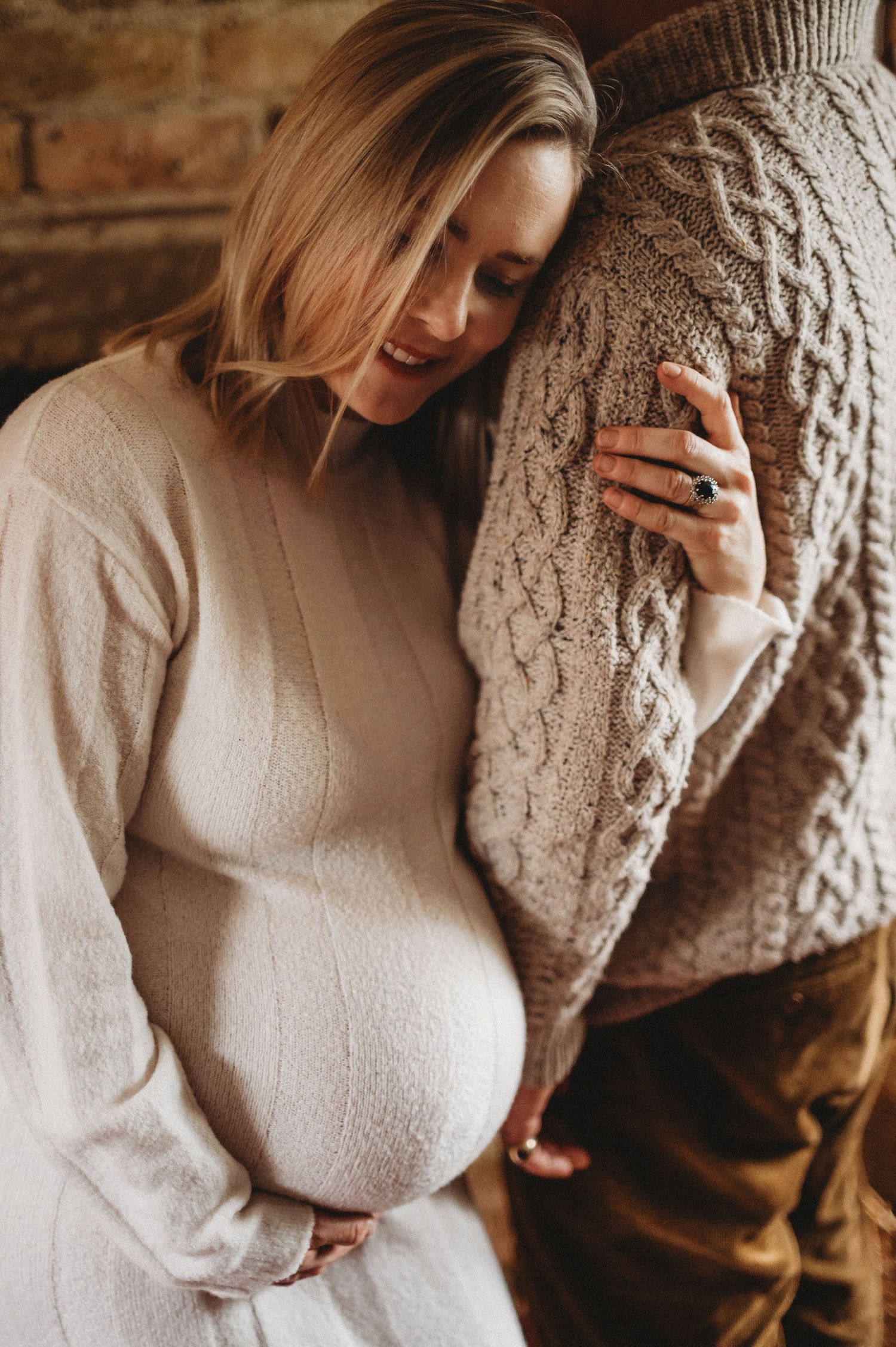Pregnant woman in cream sweater leaning against partner smiling warmly during an intimate maternity session.