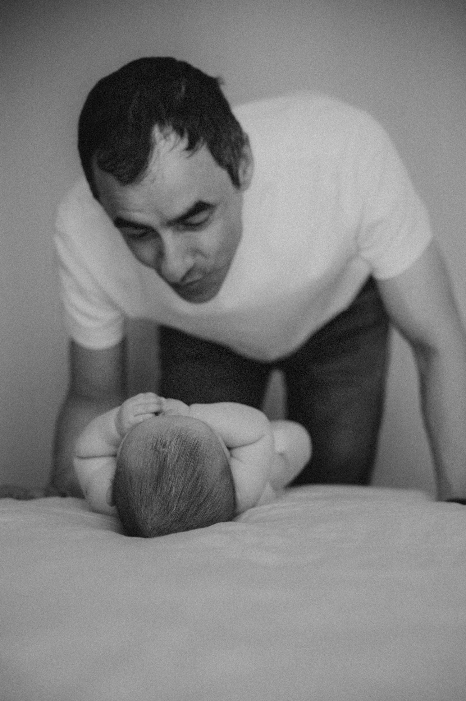 A father tenderly looks down at his newborn baby lying on a bed in black and white photo.
