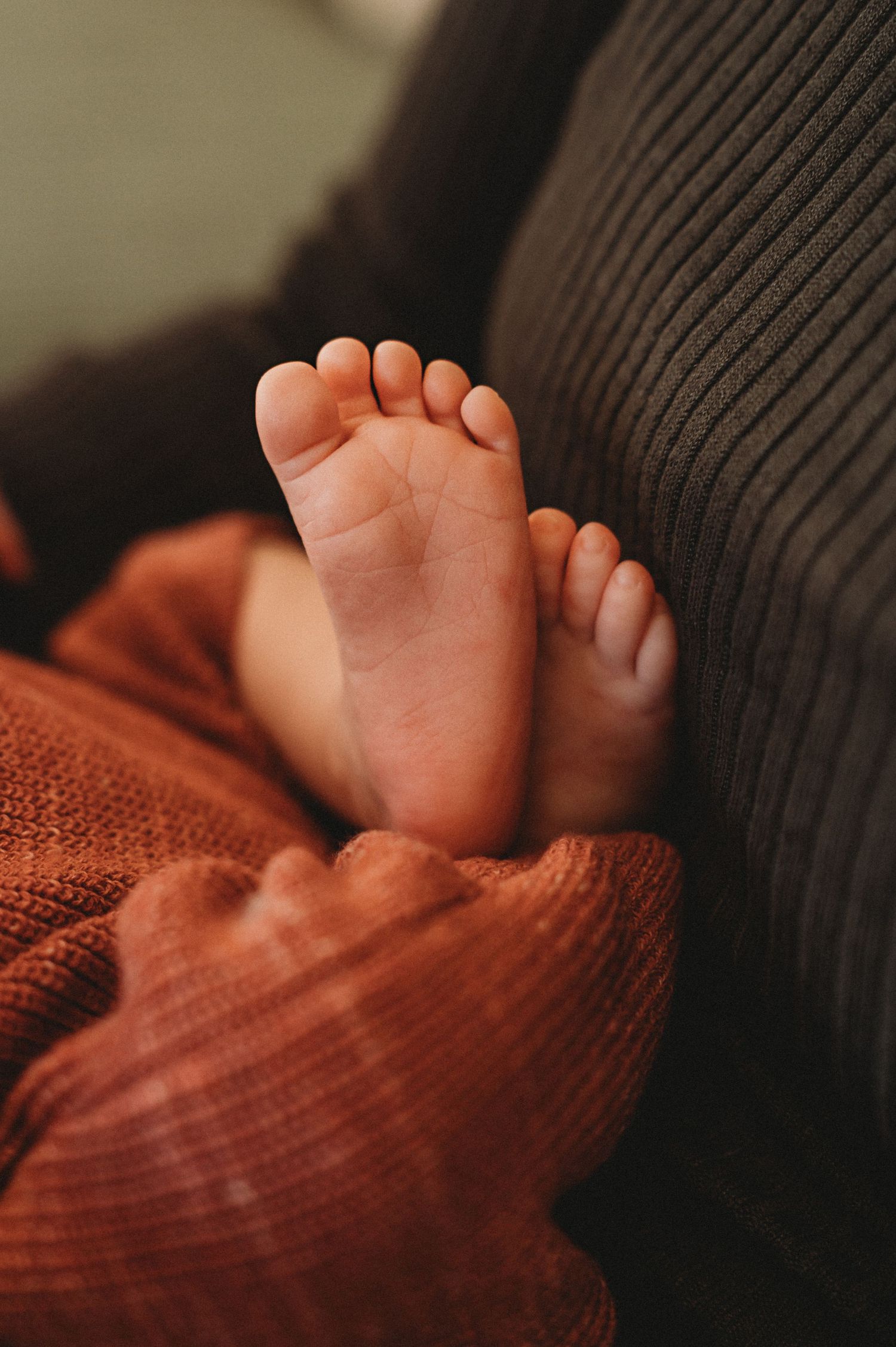 Close-up of tiny newborn baby feet peeking out from a rust-colored knit blanket on a sofa.