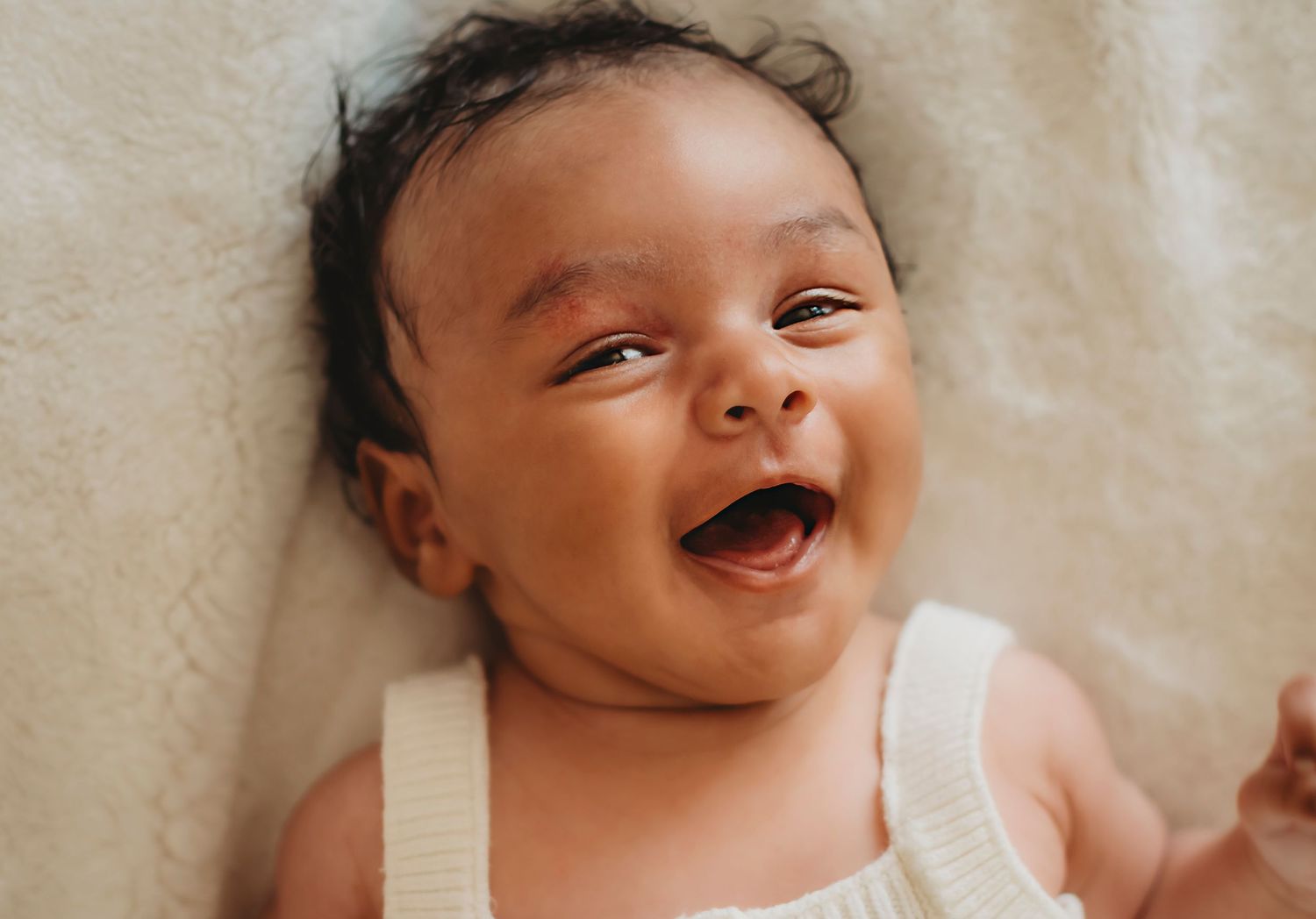 Adorable smiling dark-haired baby laughing joyfully while lying on a soft cream blanket.