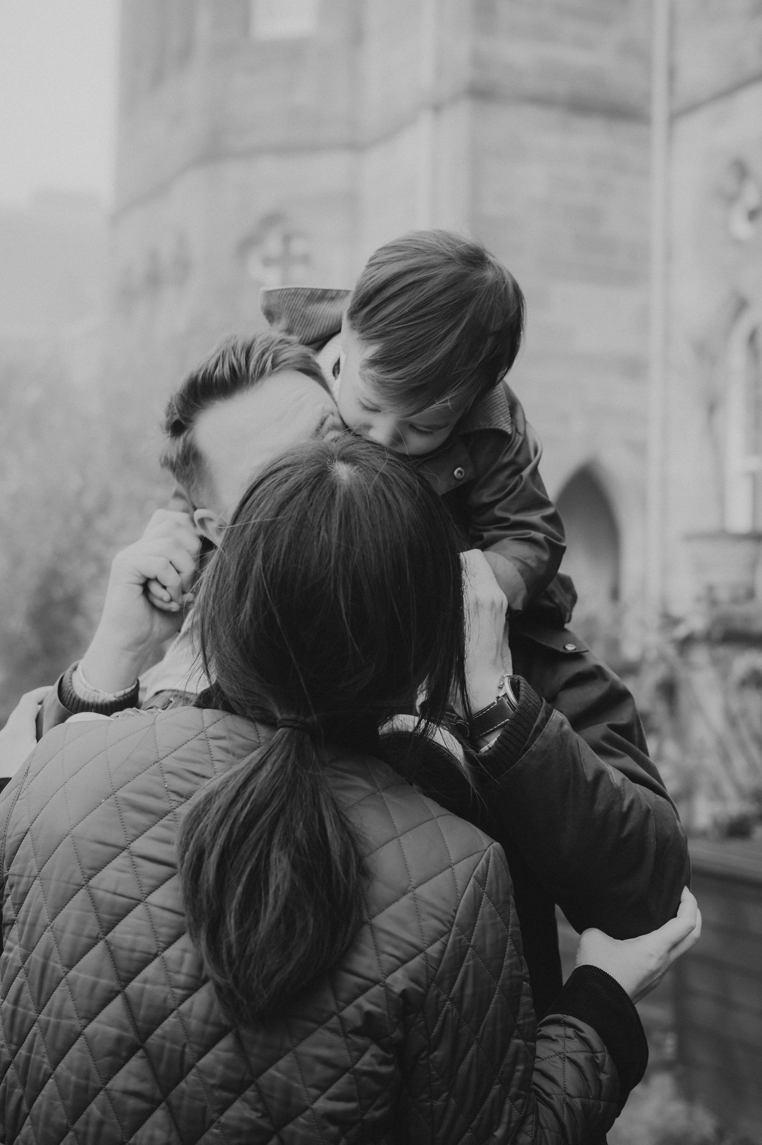 Black and white photo of parents lovingly lifting their toddler between them near a historic building.