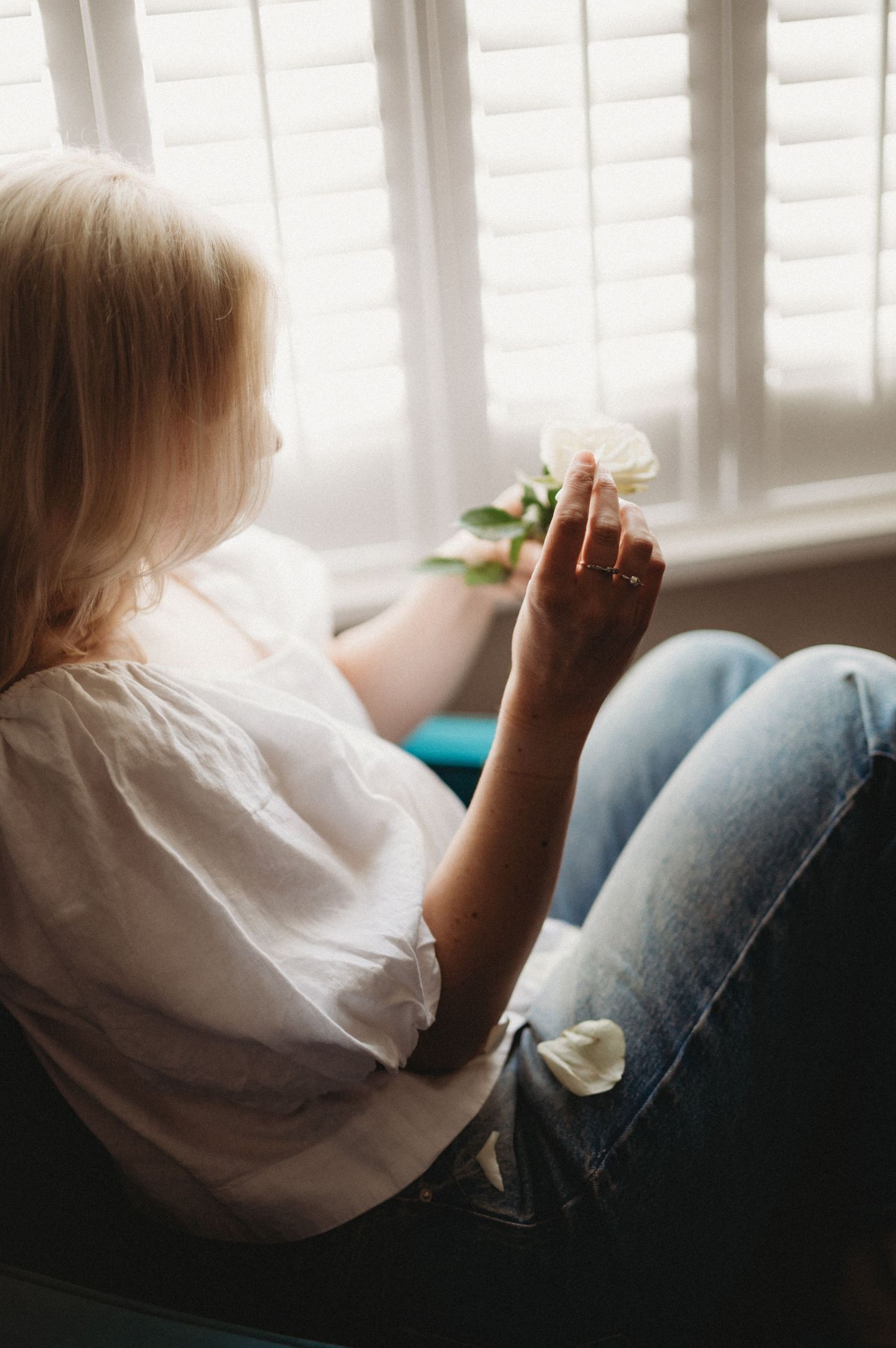 Blonde woman in white shirt and jeans holding a white flower by sunny window.