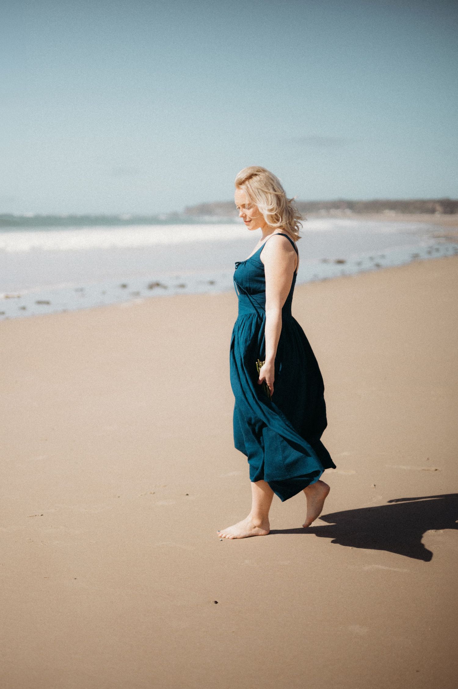 Blonde woman in teal maxi dress walking barefoot on sandy beach with ocean waves.