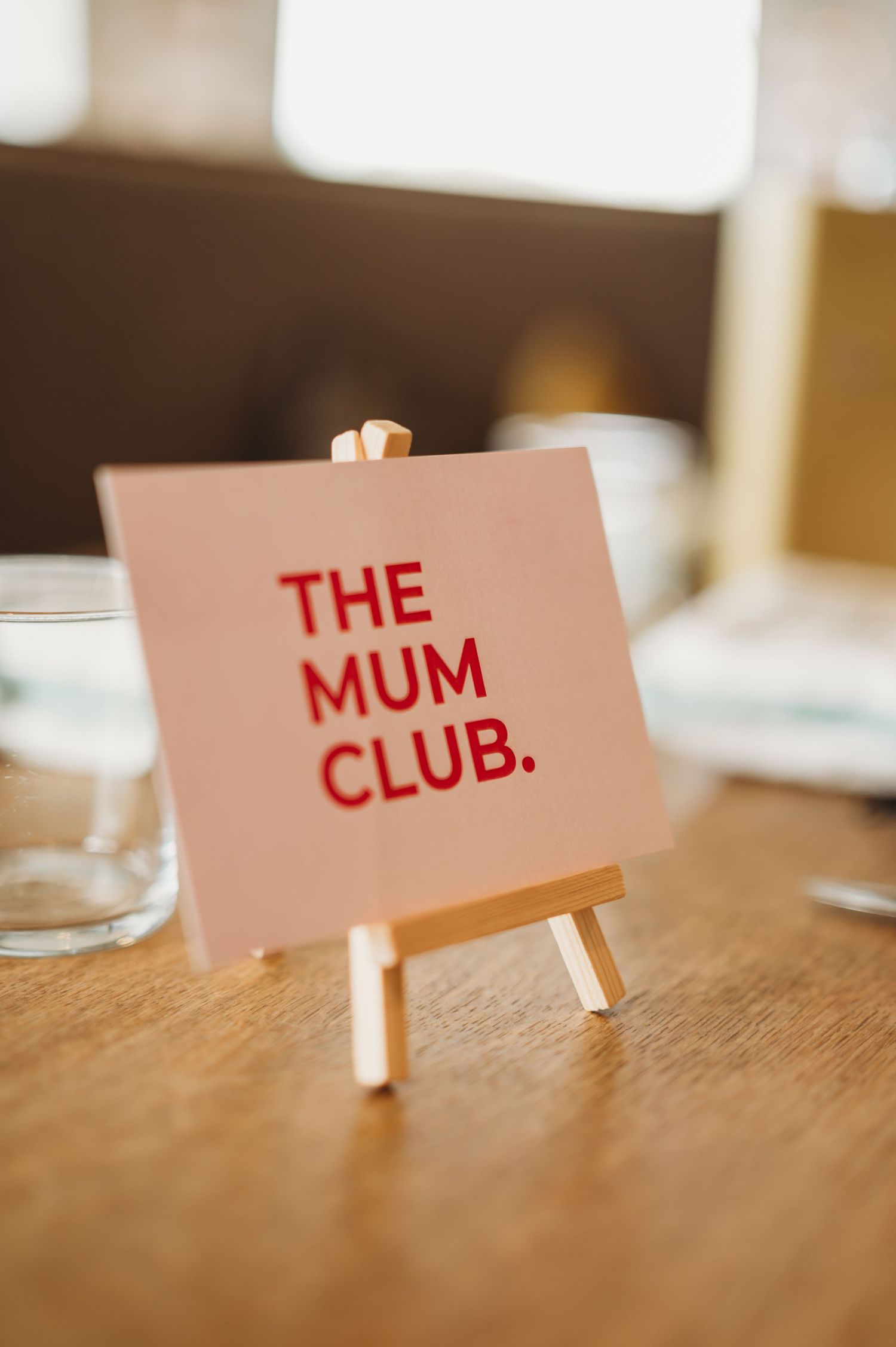 Pink card reading The Mum Club in red letters displayed on a small wooden easel on a table.