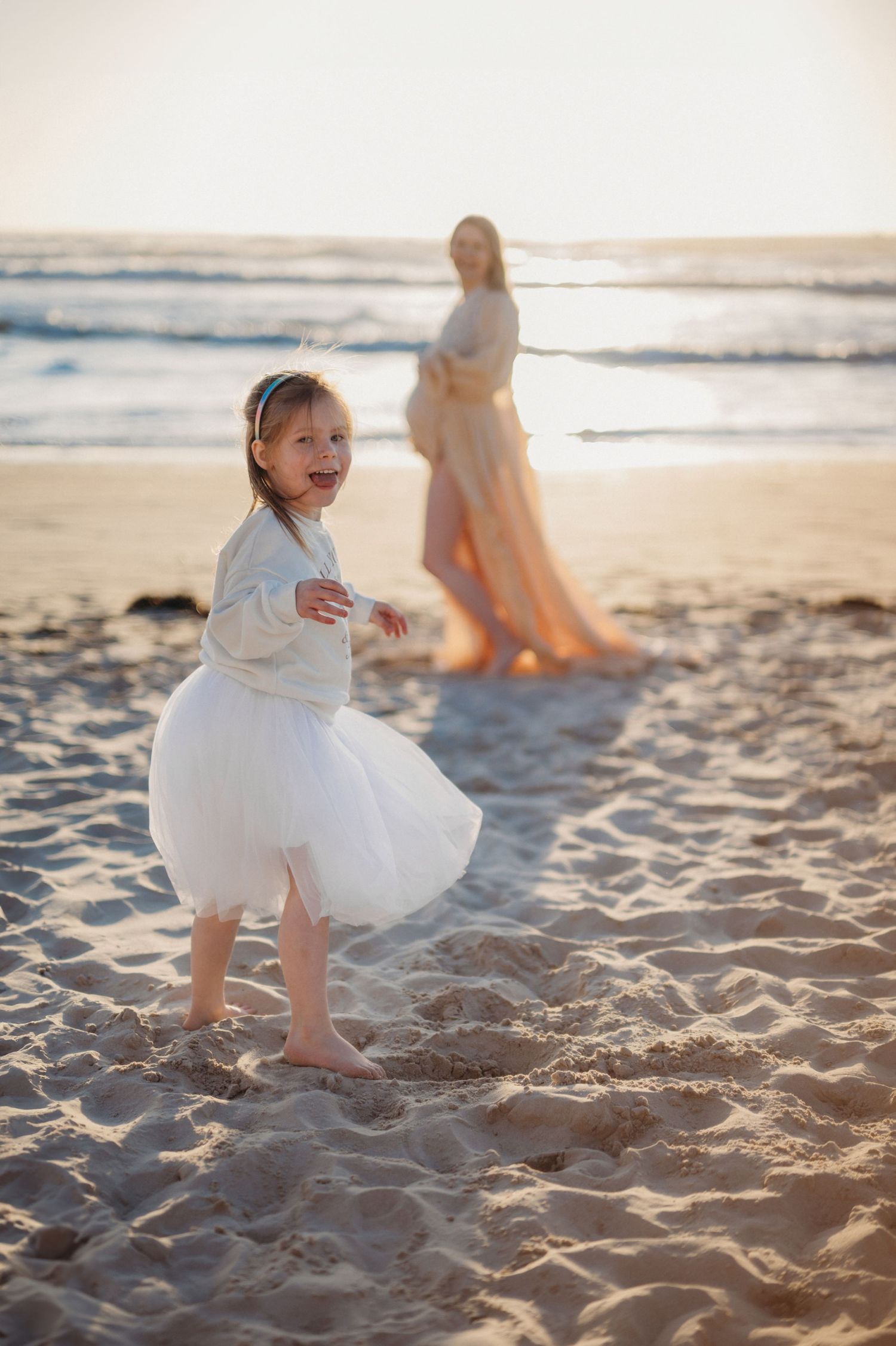 Joyful little girl in white tutu dress dancing on beach with pregnant mother standing in background.