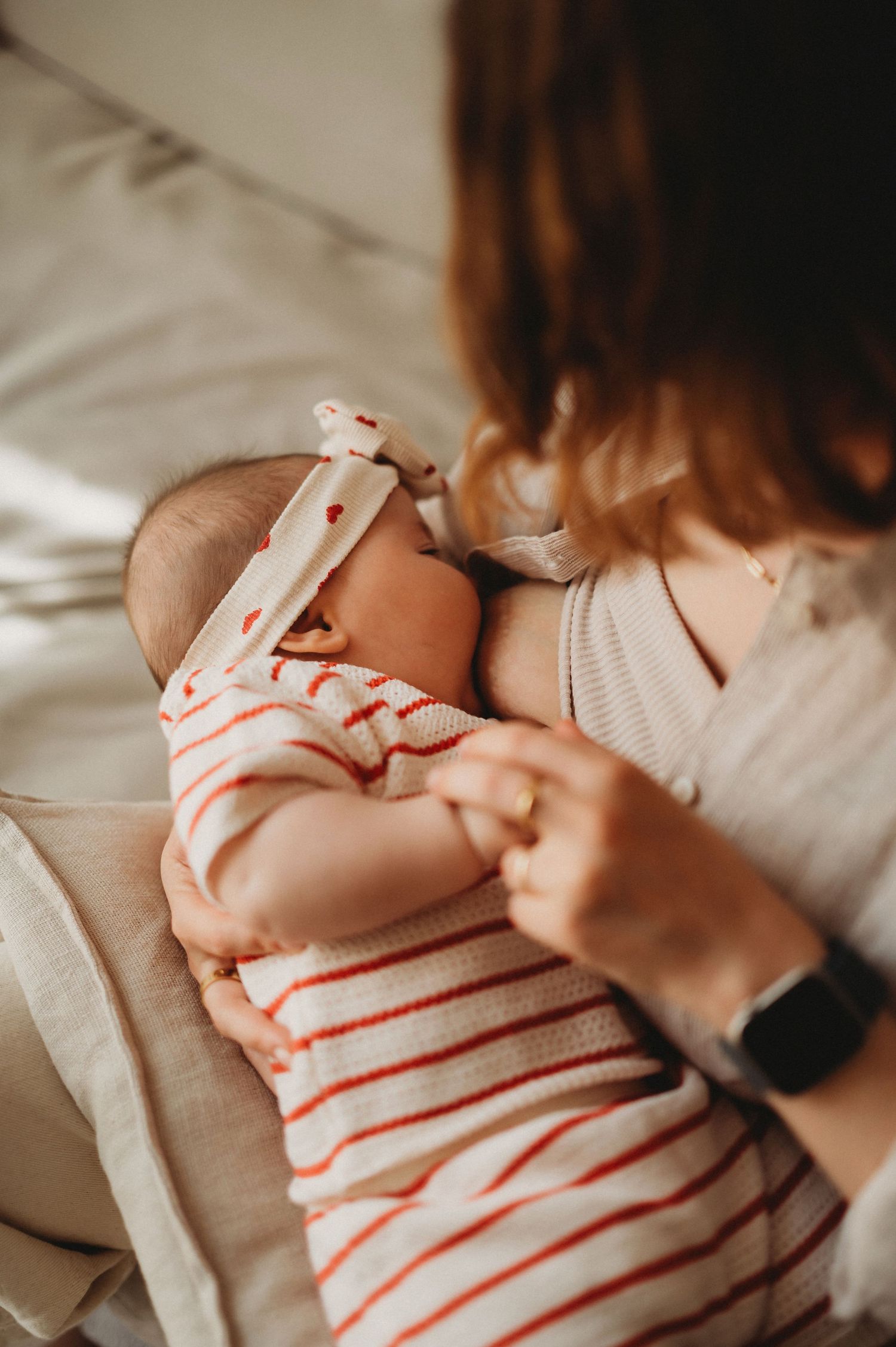 Mother gently holding sleeping newborn baby dressed in red striped onesie with bow headband.