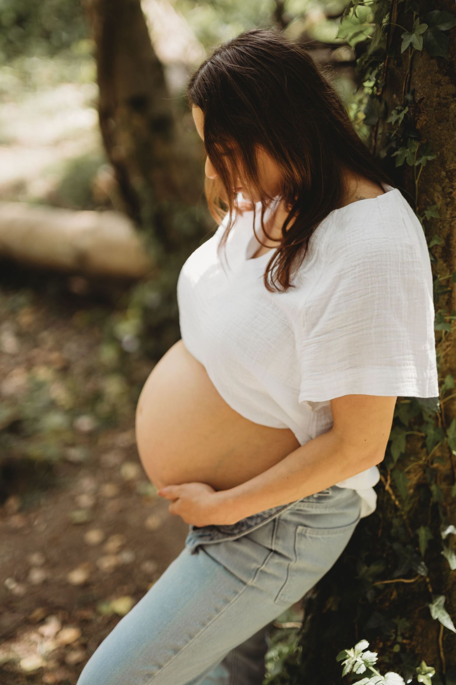 Pregnant woman in white shirt leaning against tree cradling bare bump.