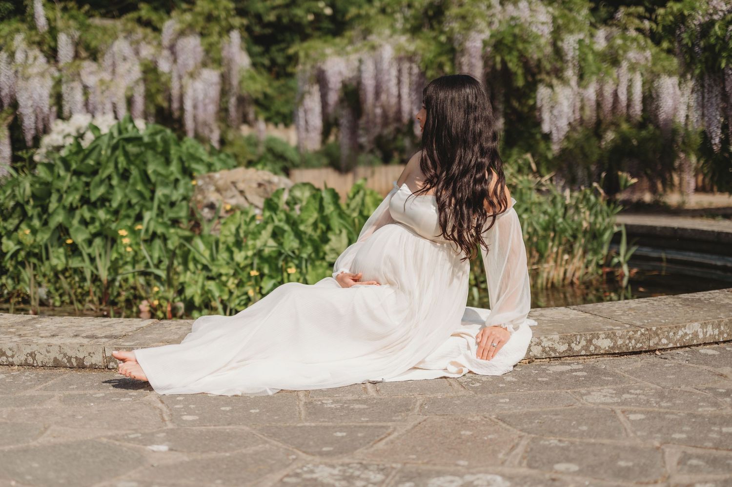 Pregnant woman in flowing white dress sitting gracefully by wisteria garden, viewed from behind.