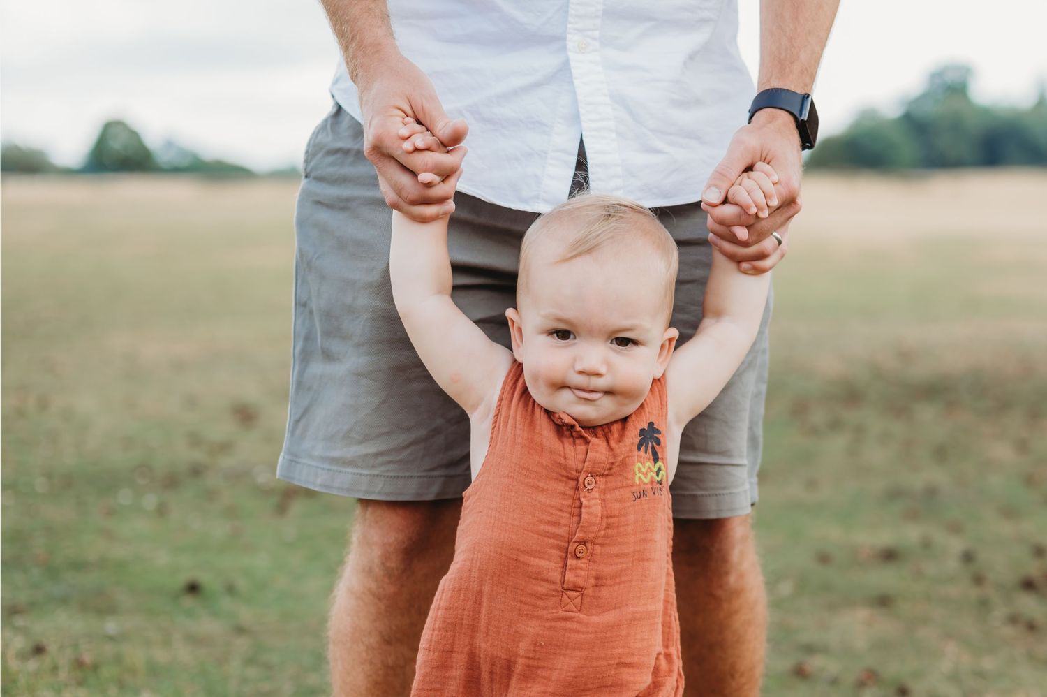 A smiling baby in an orange romper takes first steps while holding a fathers hands outdoors.
