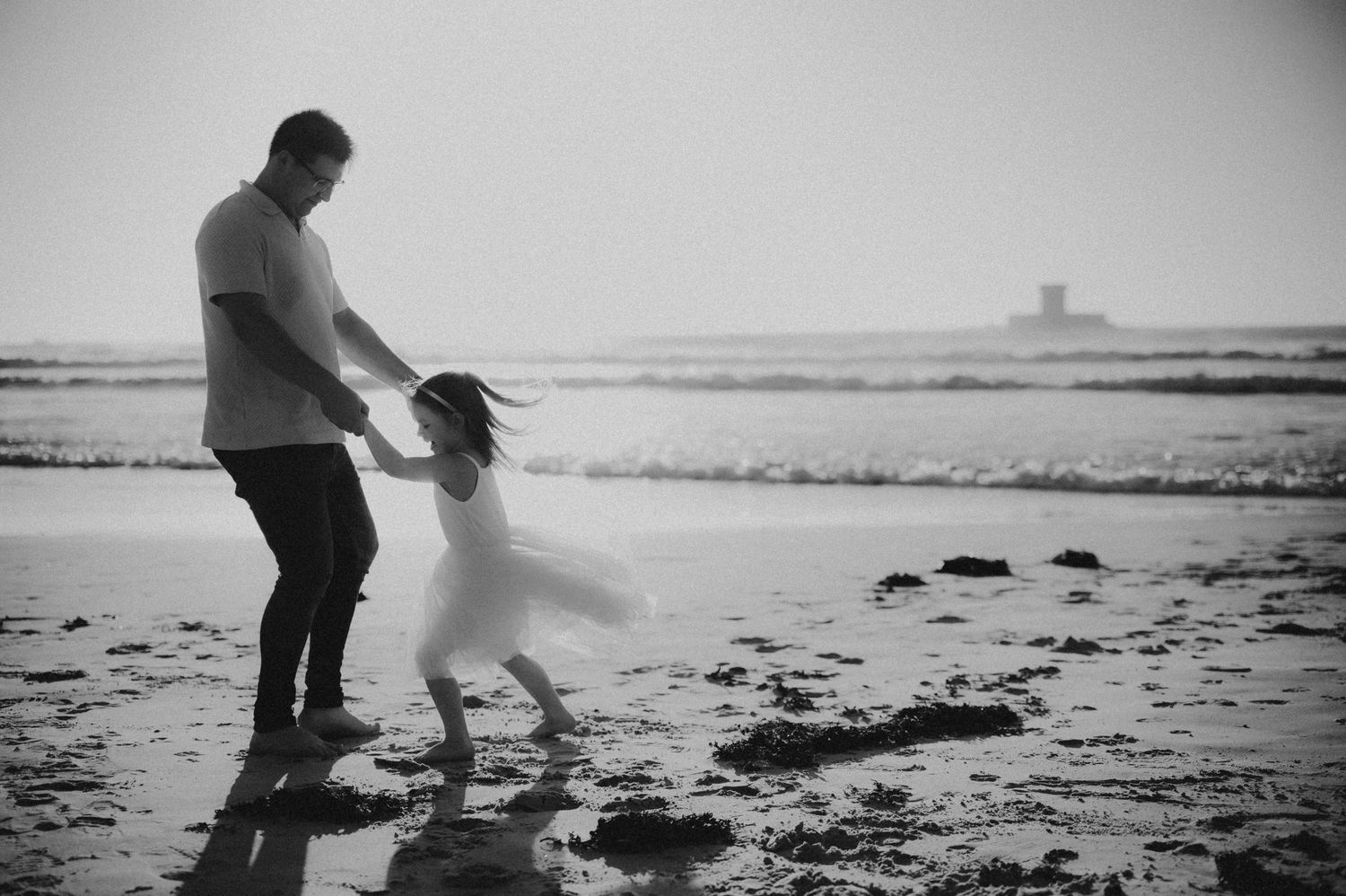 Black and white photo of father spinning daughter on beach at sunset.