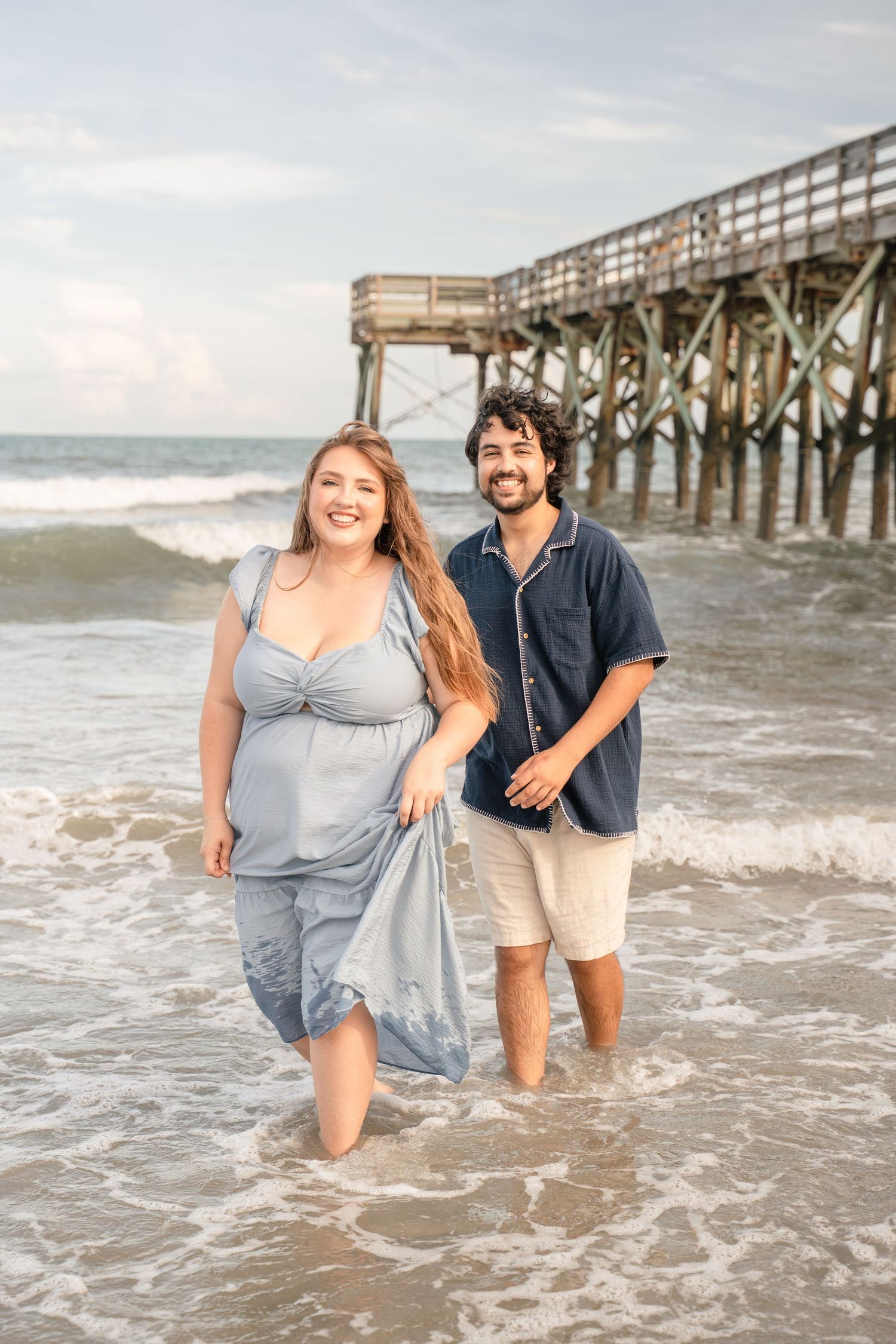 A smiling couple wading in ocean waves near a wooden pier, woman in blue dress, man in dark shirt and khaki shorts.