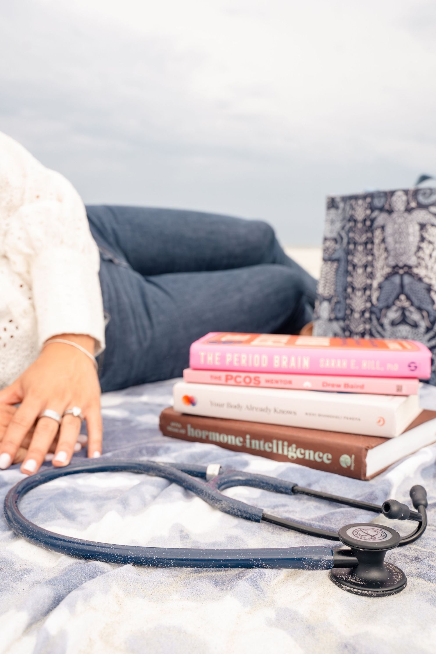 Stethoscope beside stacked women's health books including Hormone Intelligence and PCOS on a blanket outdoors.