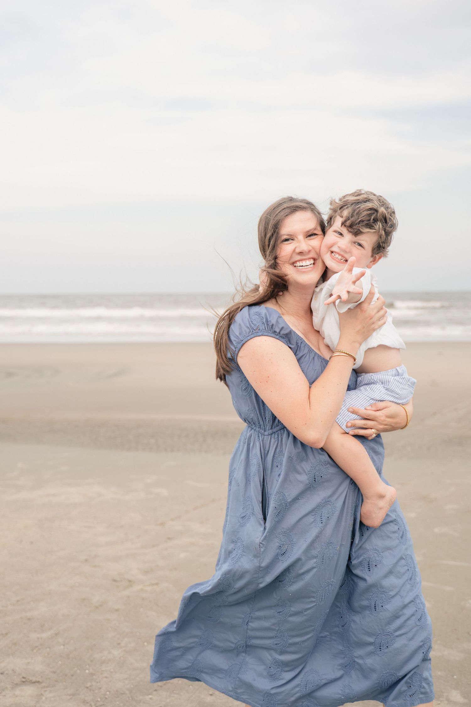 A smiling woman in a blue dress holds a young child on a sandy beach with ocean waves in the background.