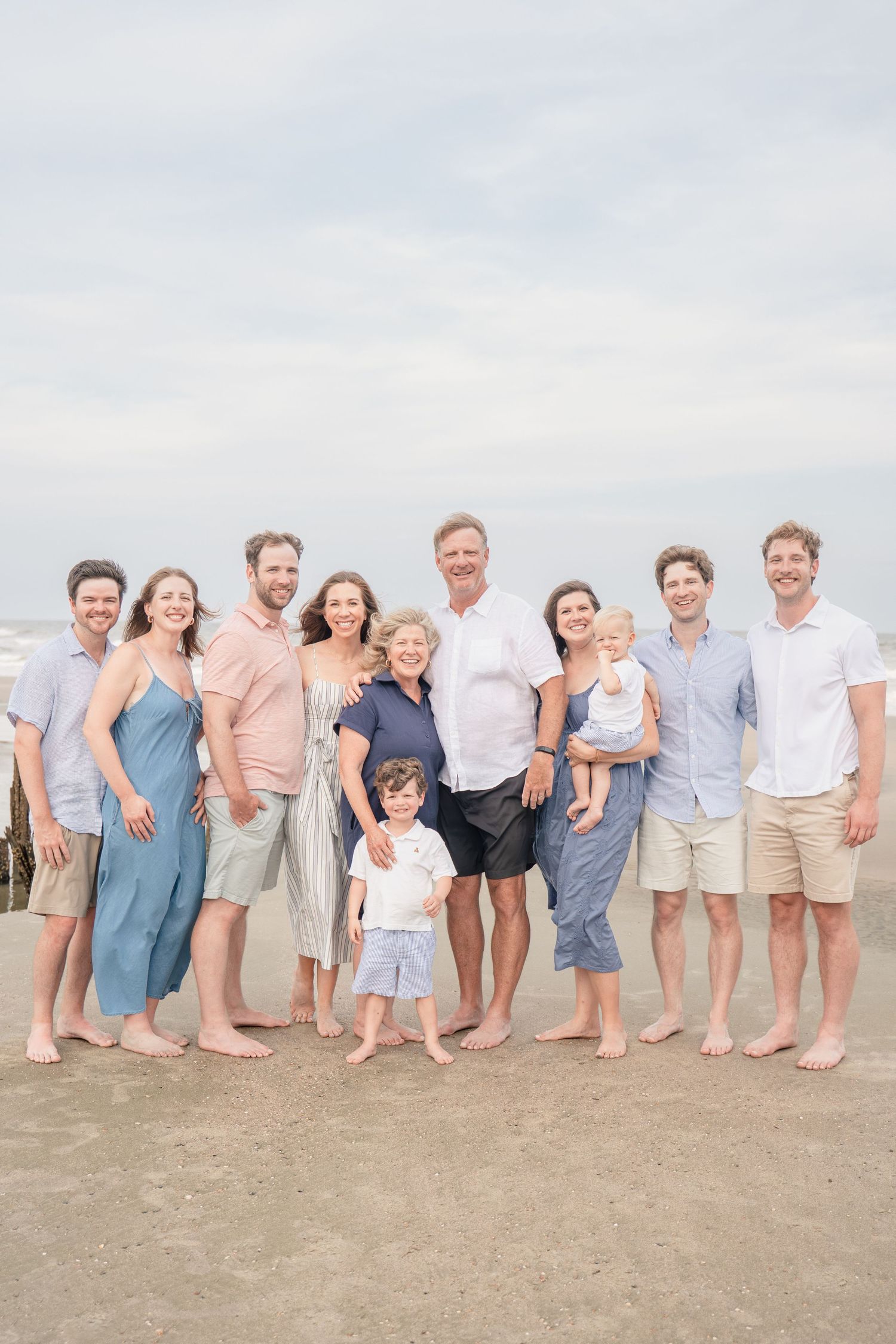 Multi-generational family of eleven poses barefoot on sandy beach, smiling together on an overcast day.