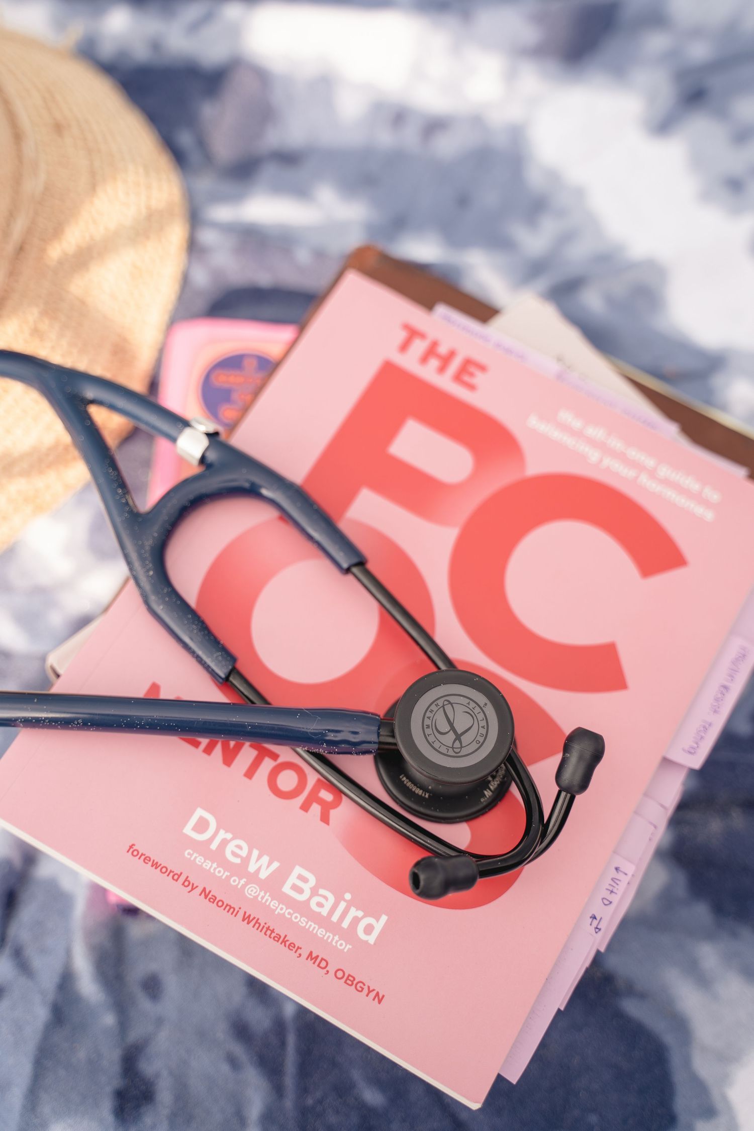 A stethoscope resting on pink medical books by Drew Baird, with a straw hat on a blue background.