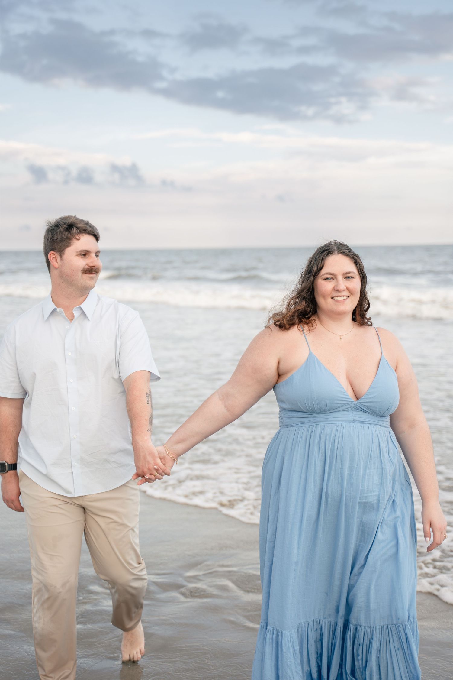 Smiling couple holding hands on beach, woman in blue maxi dress and man in white shirt walking along the shoreline.