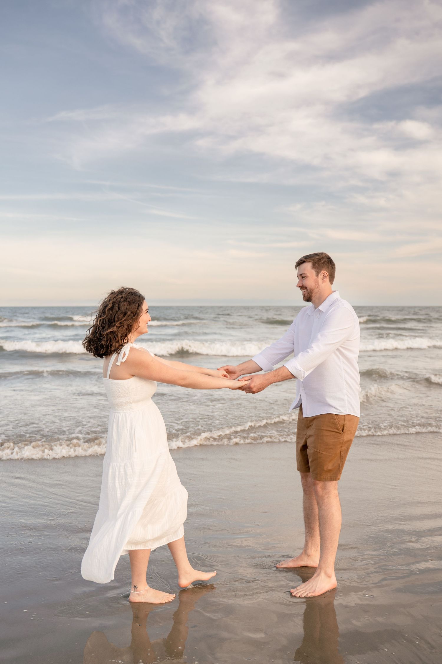 Couple holding hands on a beach at sunset, woman in white dress, man in white shirt and brown shorts, ocean waves behind.