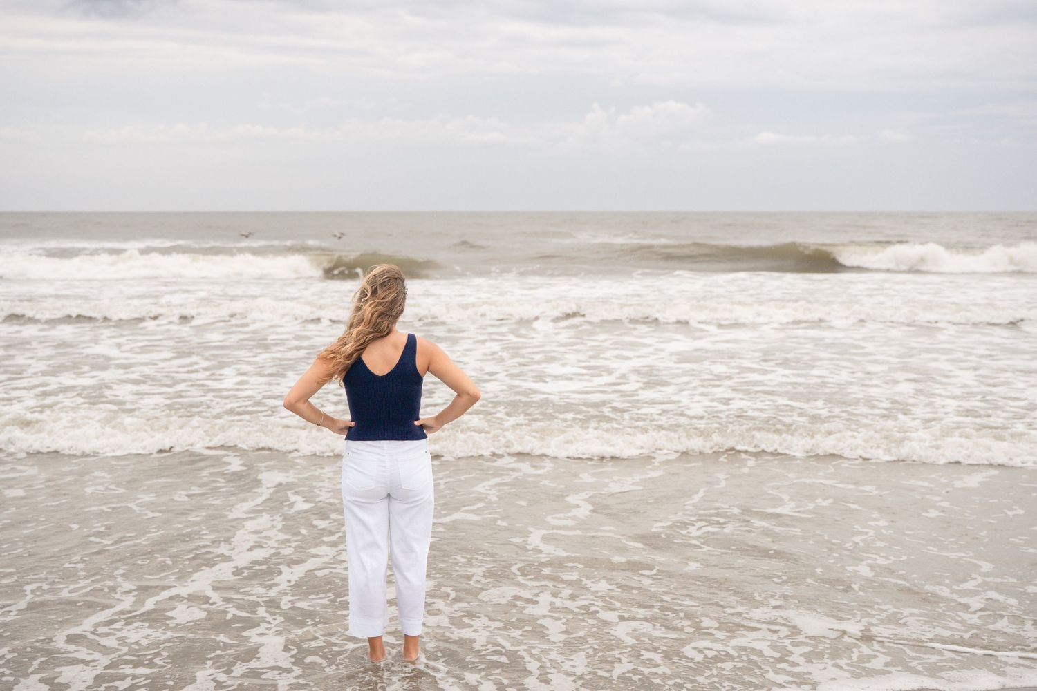 Woman in white pants and navy top stands at ocean's edge, waves washing ashore under overcast sky.
