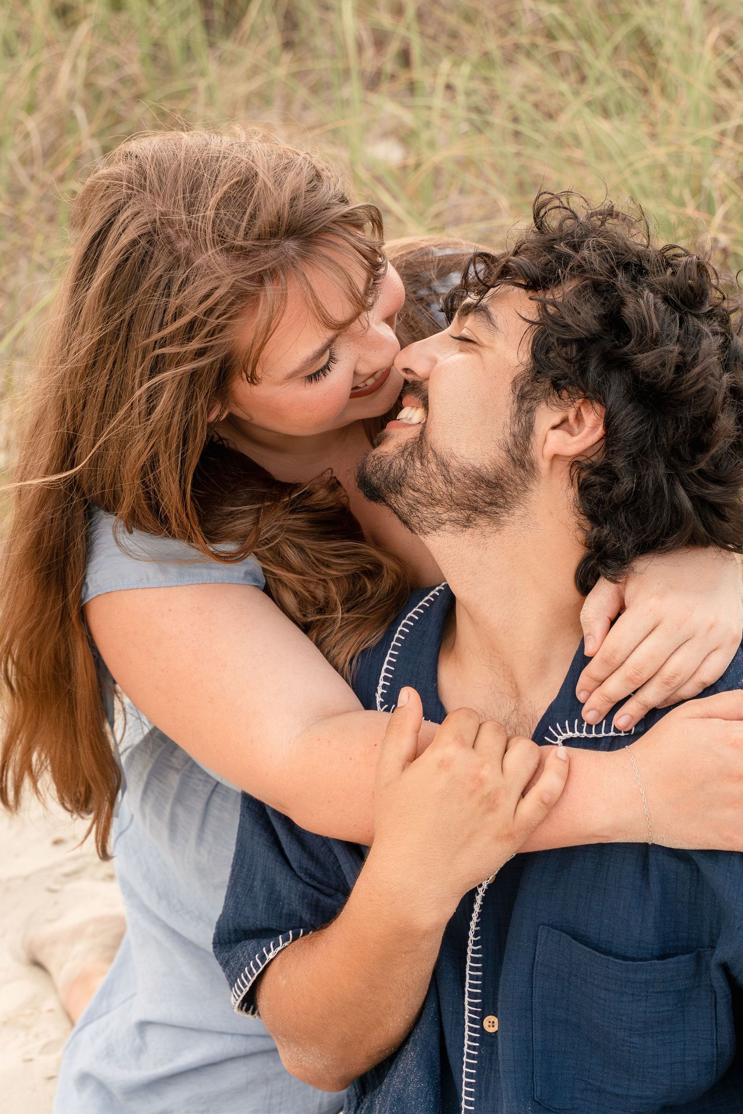 A happy couple embracing and sharing a romantic moment outdoors on a sandy beach with tall grass in the background.